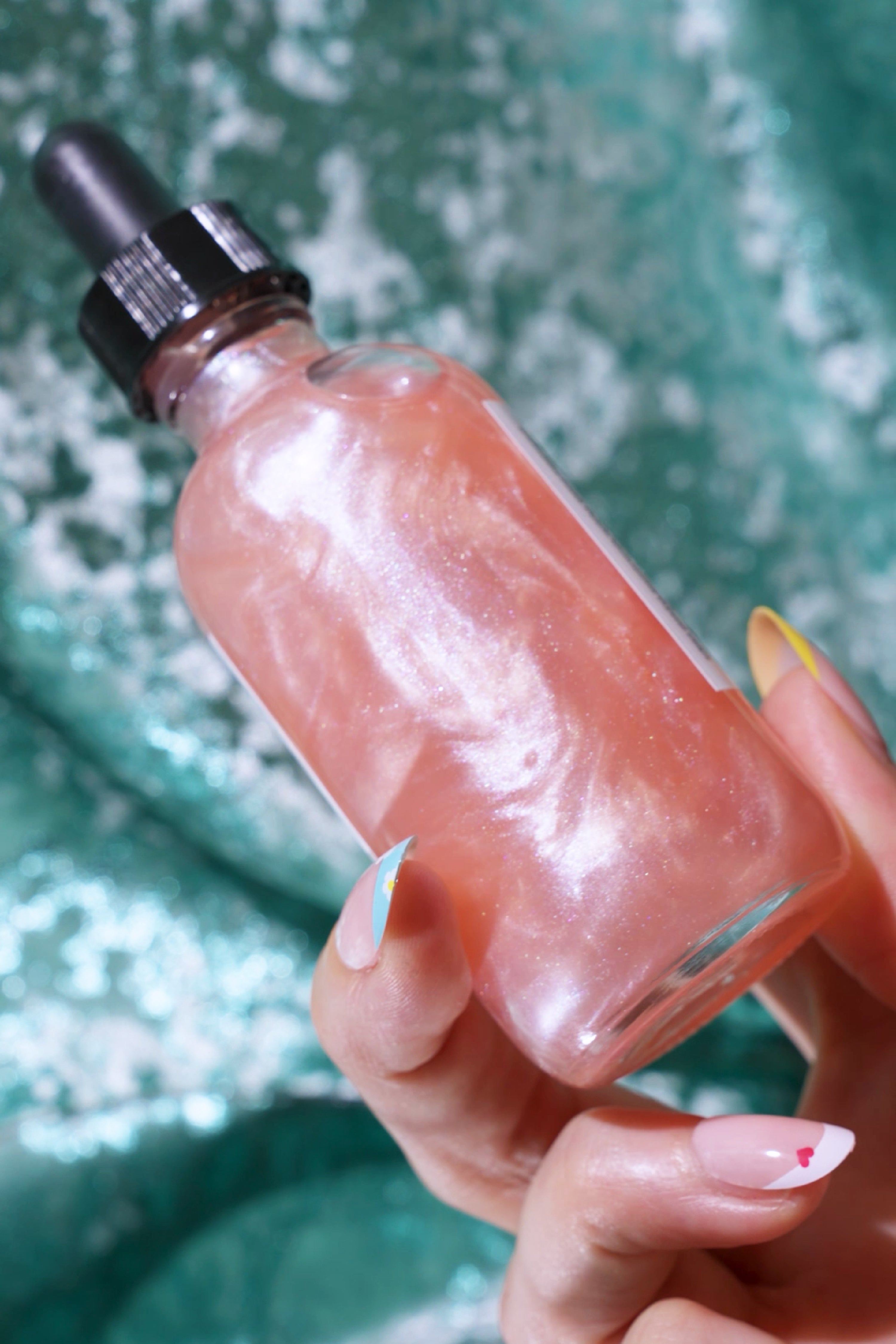 Hand holding a pearlescent pink bottle of Adventures Await Bath & Body Oil with a black dropper cap, shimmering against a sparkling green textured background.