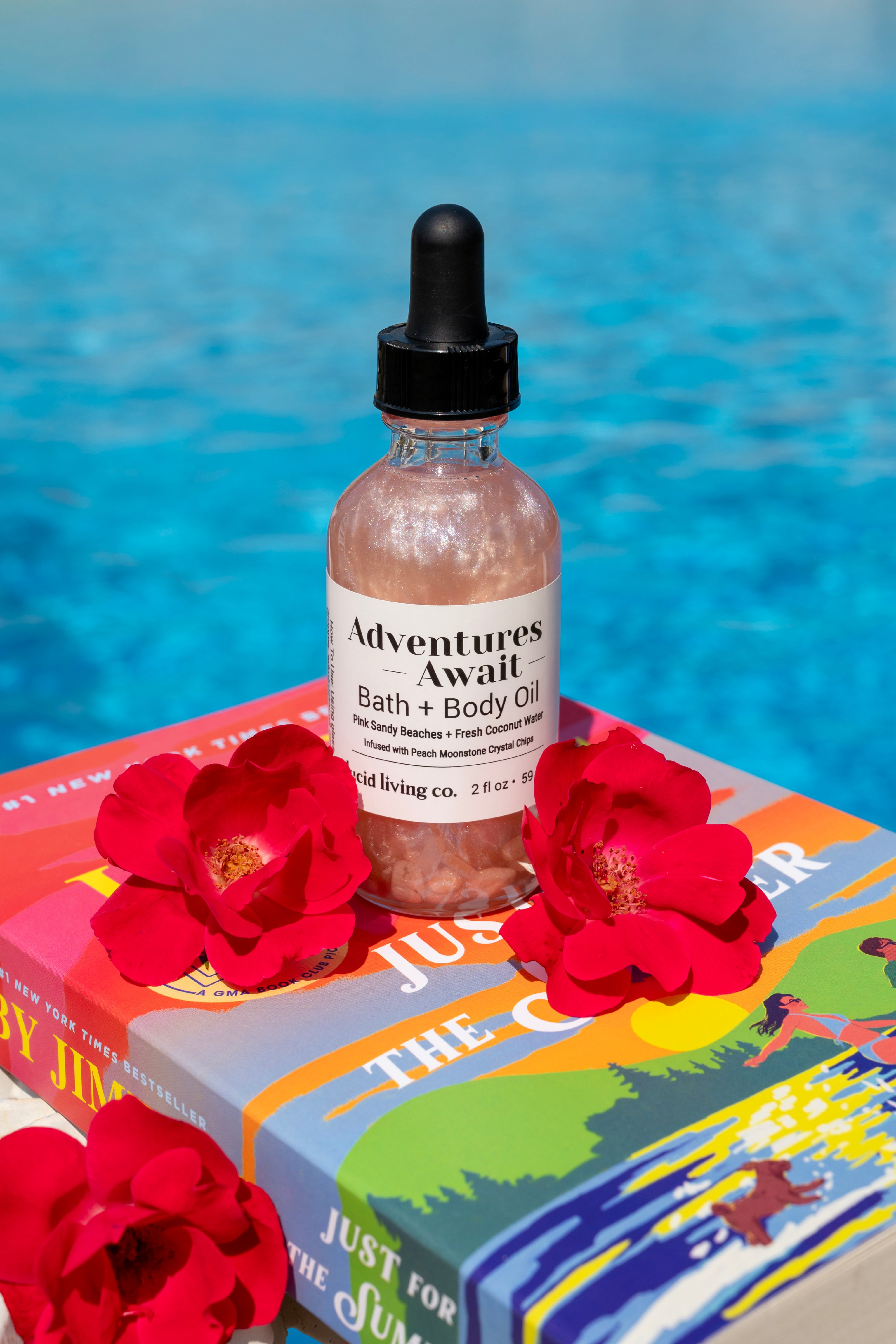 Clear glass bottle of Adventures Await Bath & Body Oil with pearlescent shimmer and black dropper cap, surrounded by red flowers, set against a vibrant book and blue water background.