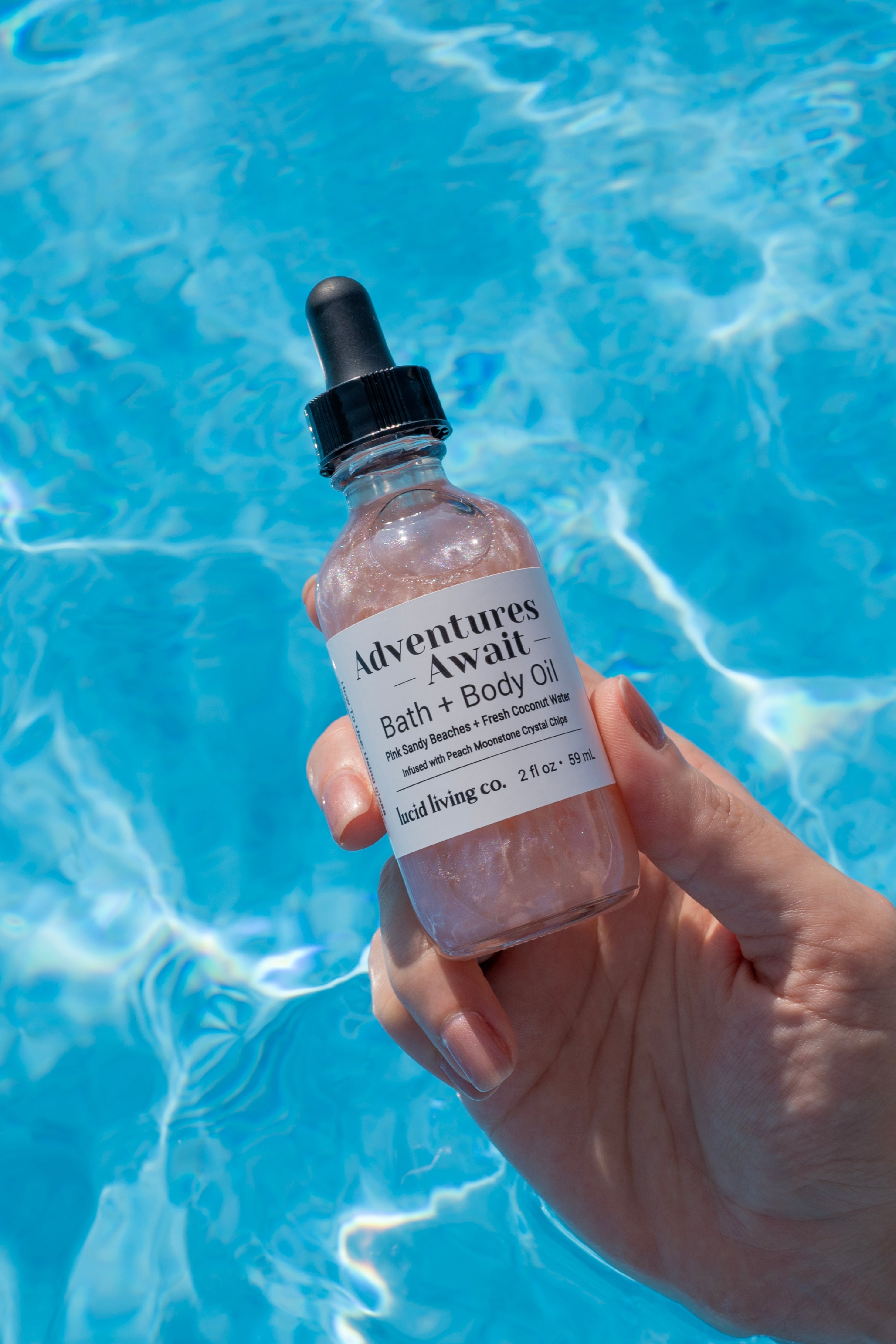 Hand holding a shimmering bottle of Adventures Await Bath & Body Oil with a black dropper cap, glowing softly above clear blue water reflecting sunlight.