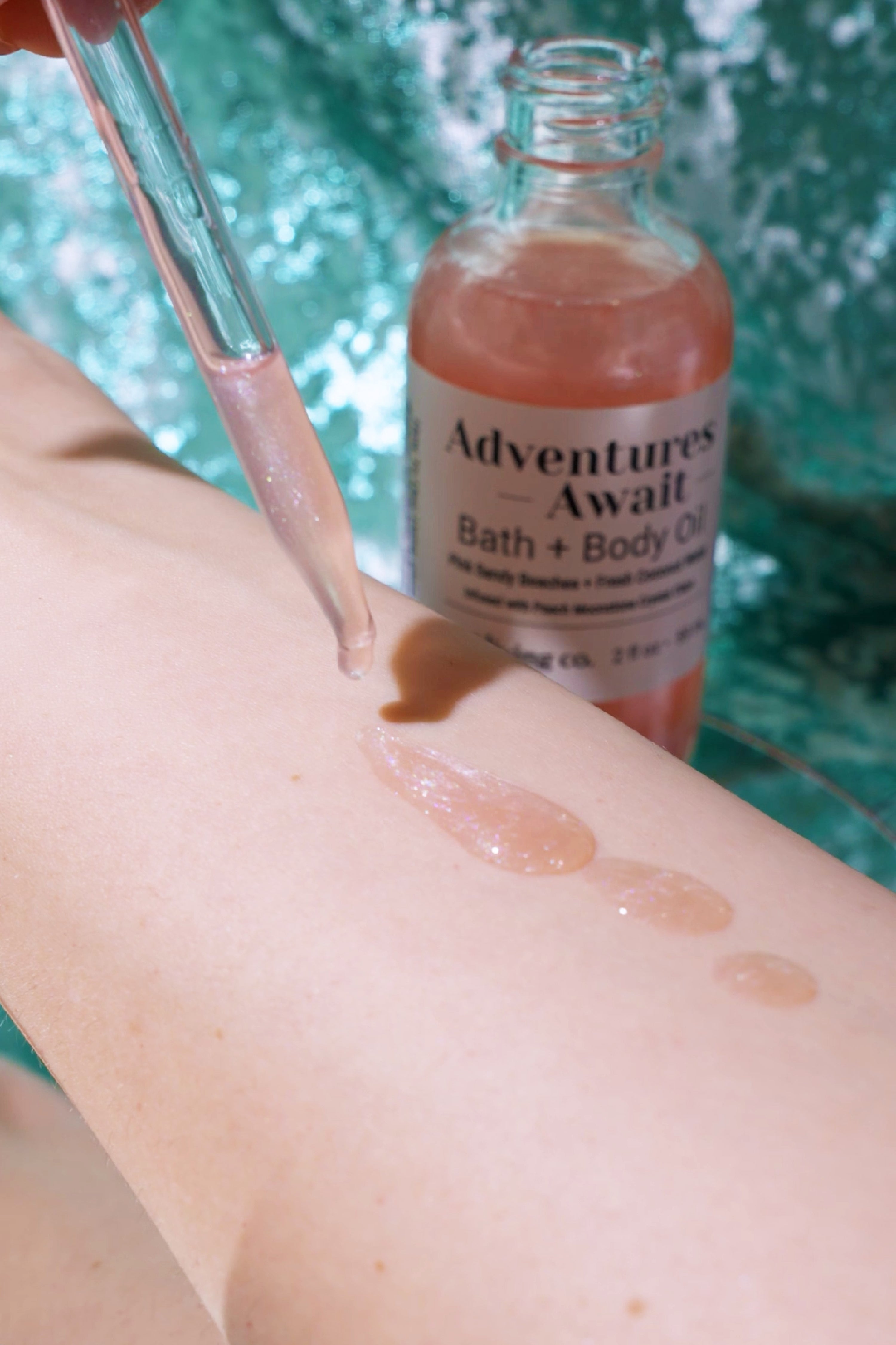 Delicate pink Adventures Await Bath & Body Oil being dripped from a glass dropper onto smooth skin, highlighting its shimmering texture with a softly blurred green background.