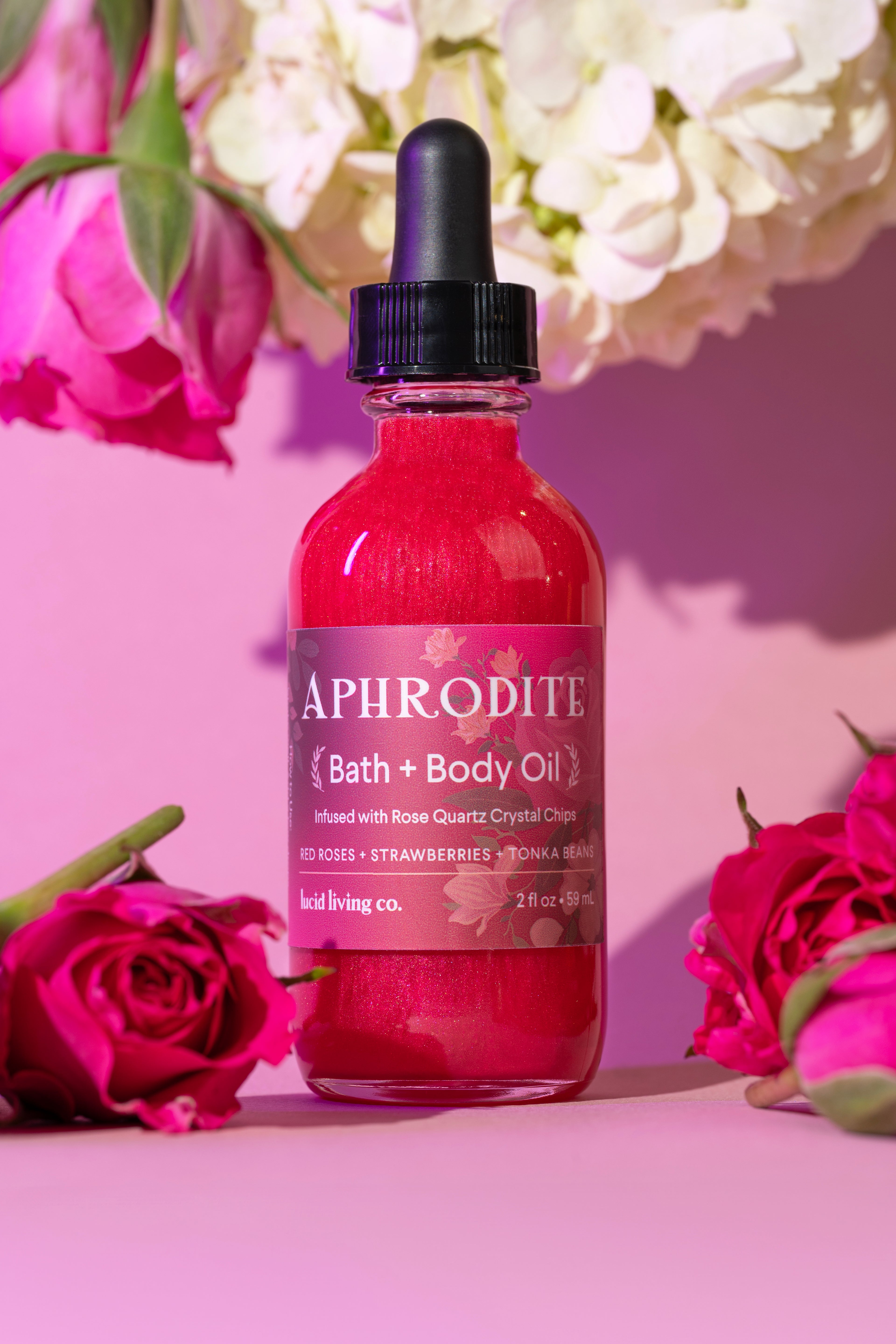 Aphrodite Bath & Body Oil bottle with rose quartz infused, surrounded by pink roses and hydrangea flowers on pink background.