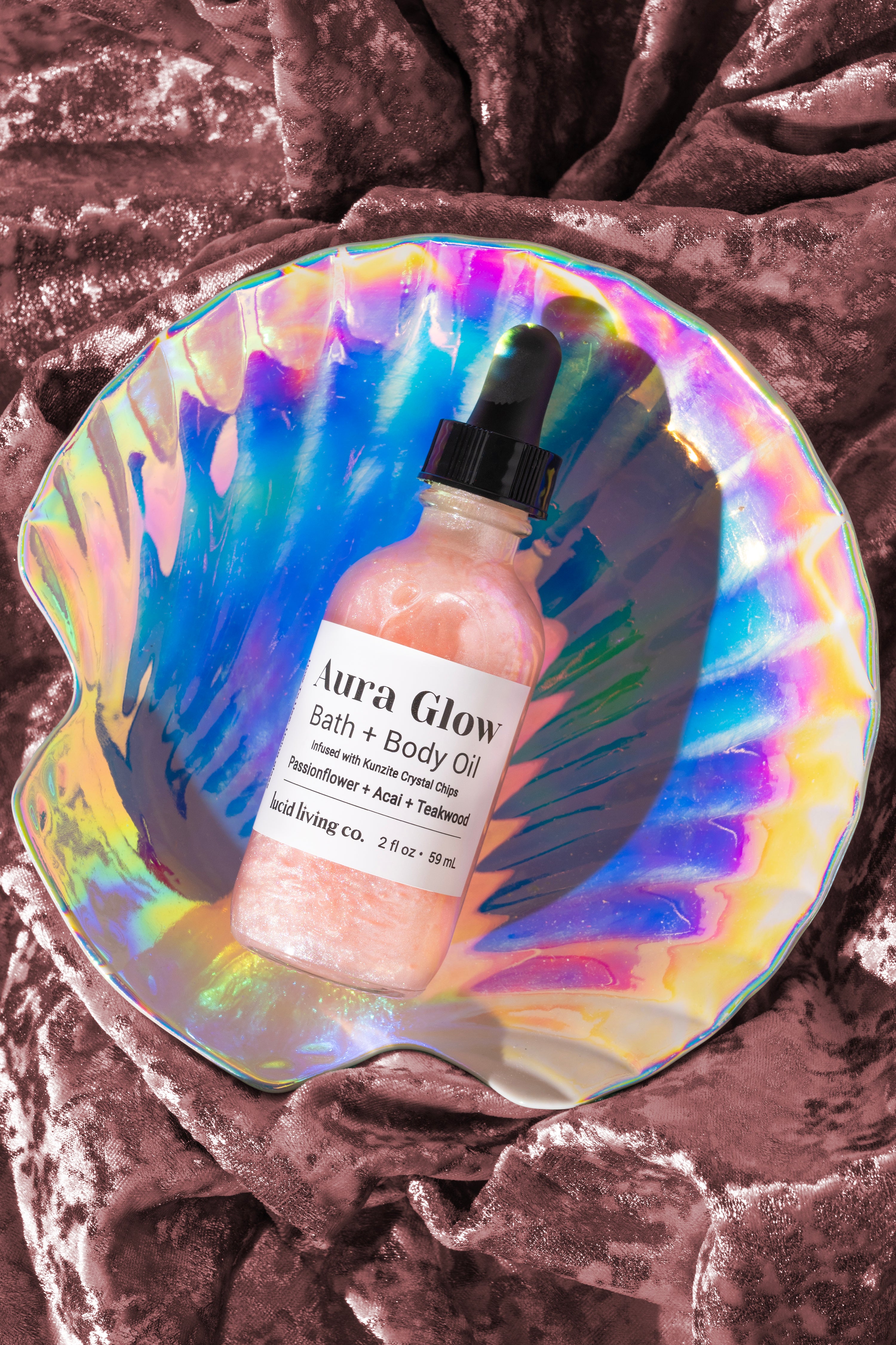 Aura Glow Bath & Body Oil in a shimmering pink liquid with visible suspended particles, resting inside a iridescent shell-shaped dish on soft textured fabric.