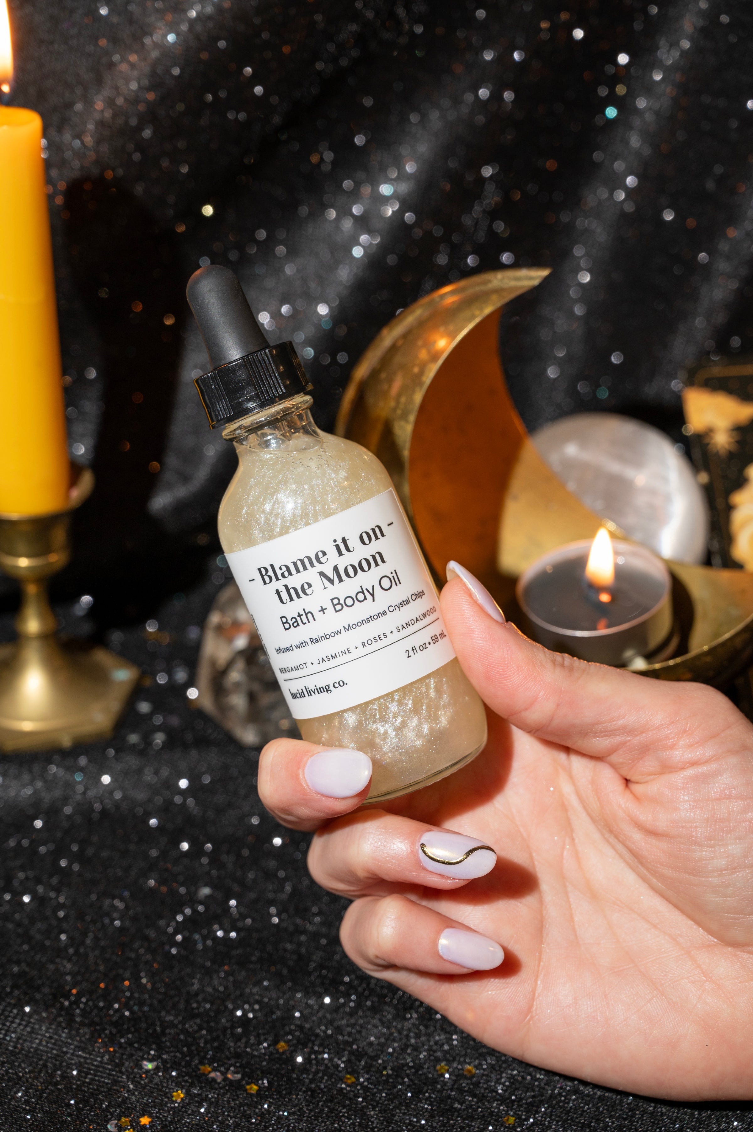 Hand holding a shimmering bottle of Blame it on the Moon Bath & Body Oil with a black dropper cap, set against a dark sparkling background with lit candles and decorative moon accents.
