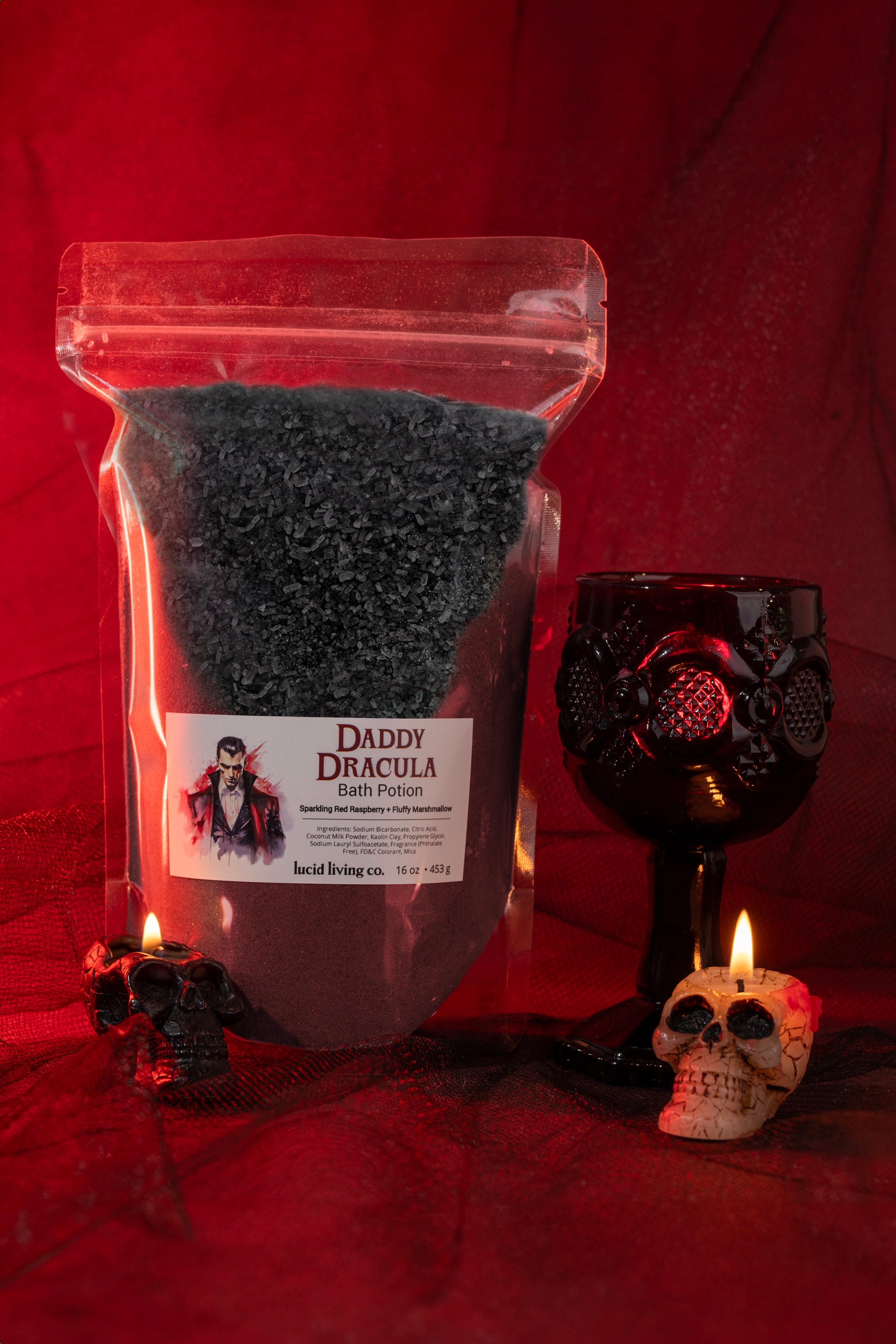 Daddy Dracula Bath Potion with black bath salts, gothic skull candles, and a dark ornamental goblet on red fabric background.