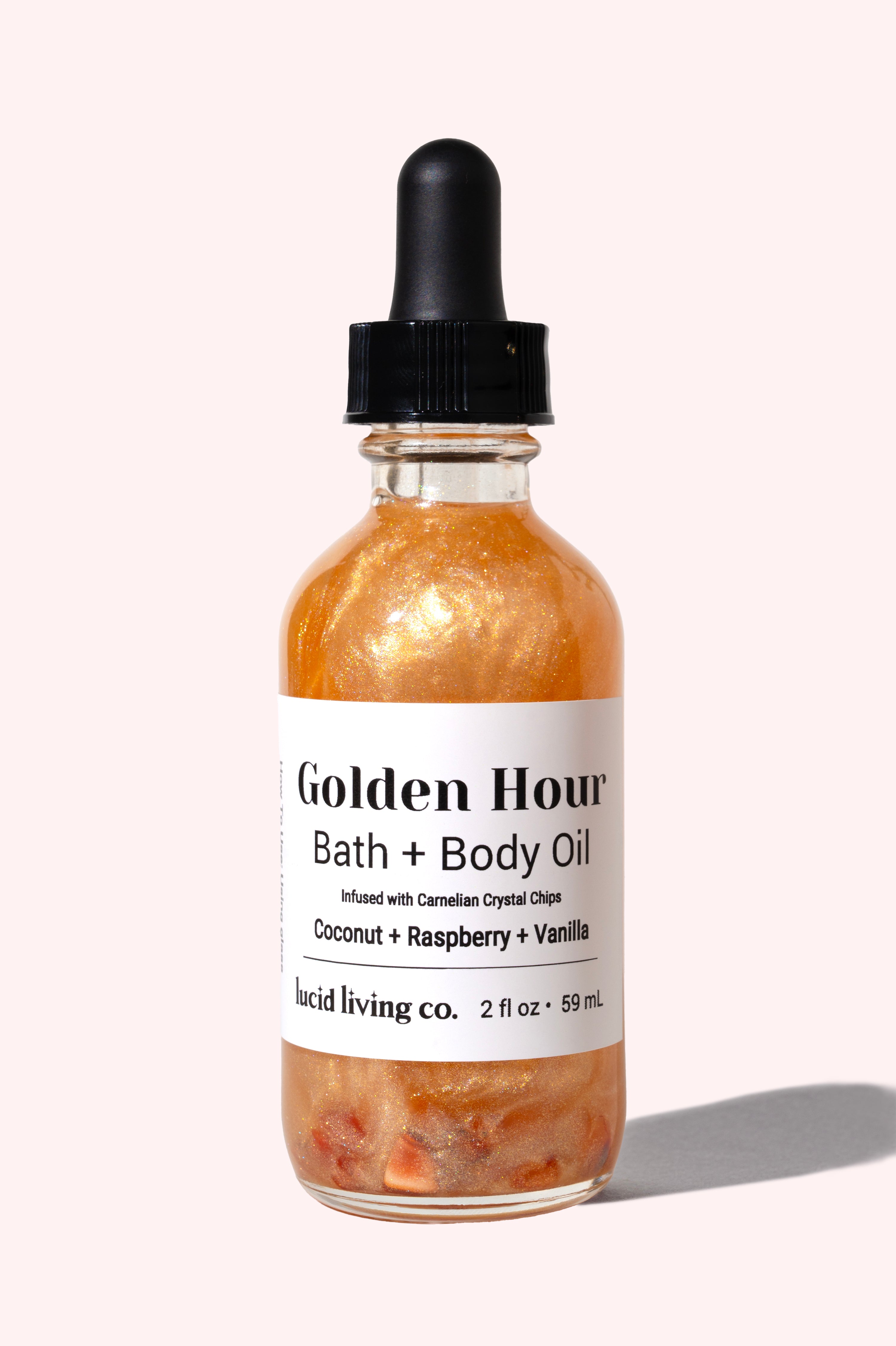 Clear glass bottle of Golden Hour Bath & Body Oil with shimmering golden liquid and visible carnelian crystal chips inside, featuring a black dropper cap on pale background.