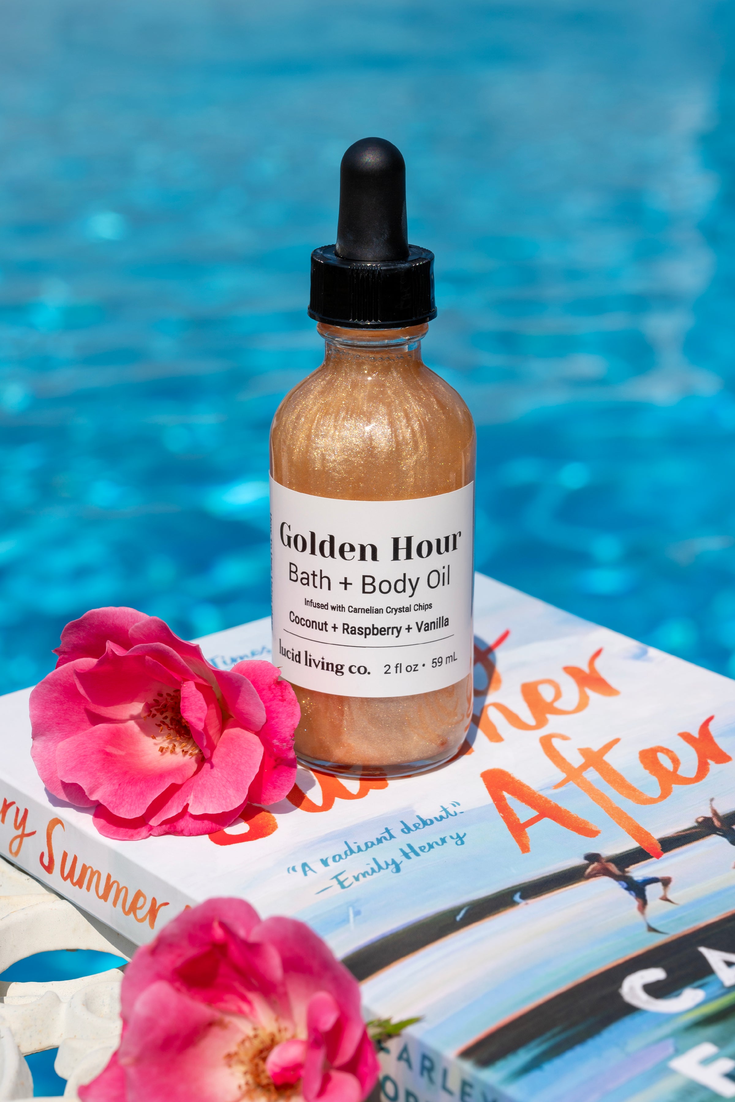 Golden Hour Bath & Body Oil in a shimmering golden liquid with black dropper, placed on a summer-themed book near pink flowers and bright blue water, evoking a radiant scent experience.