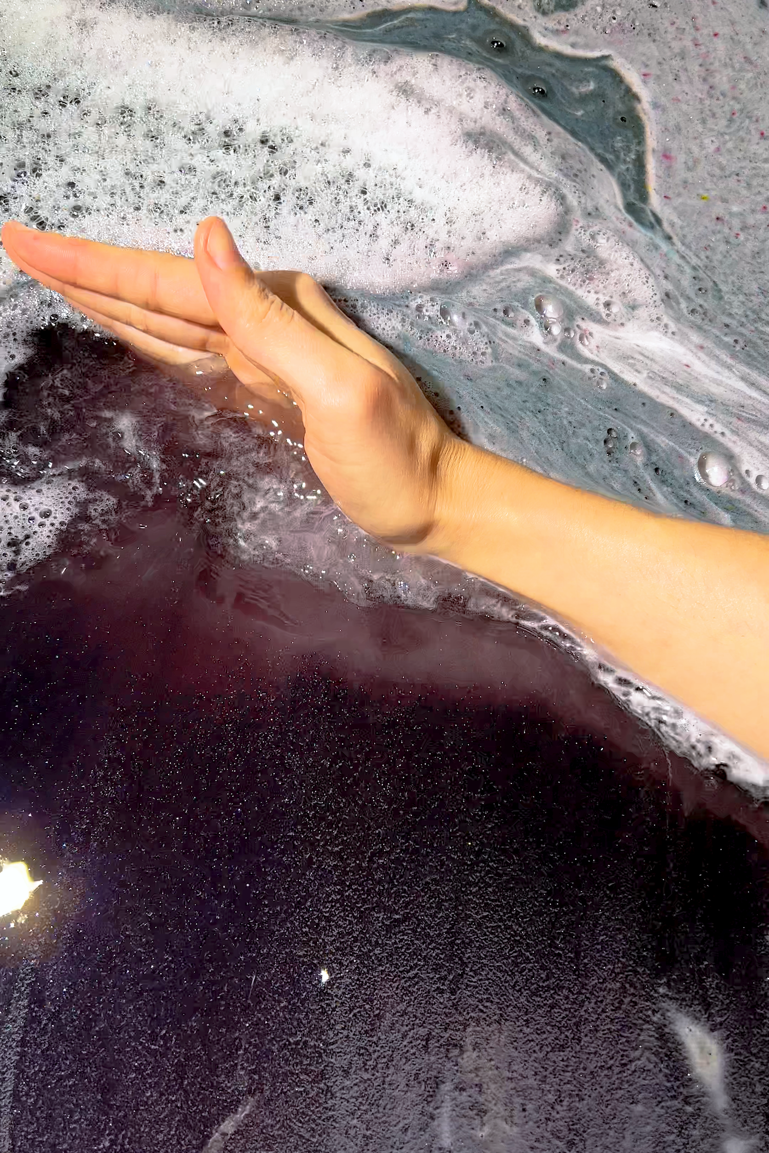 Hand stirring dark foamy water showing bubbles and colors dissolving for LILITH "The Dark Mother" Bath Potion.