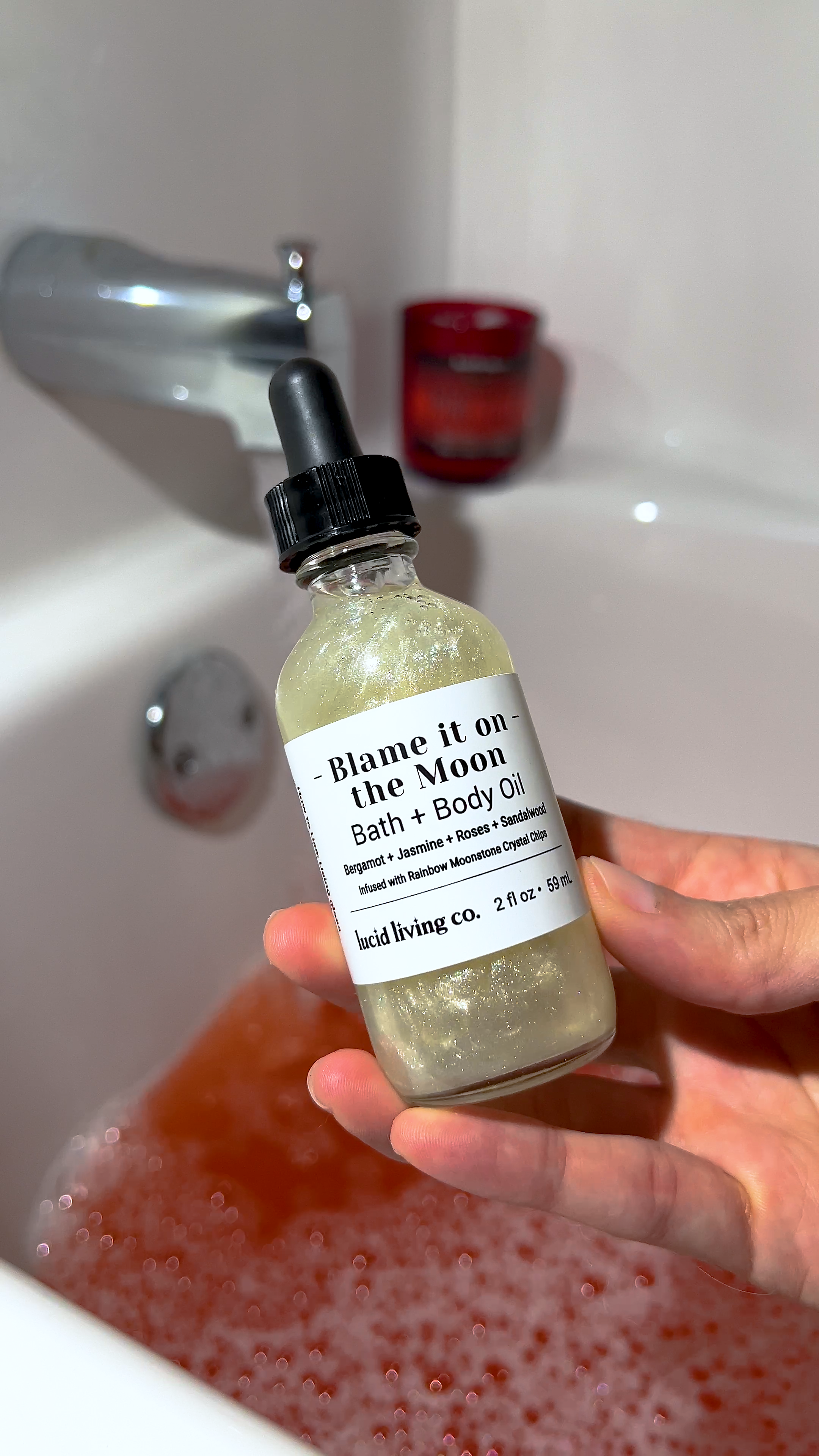 Blame it on the Moon Bath & Body Oil held over a bath filled with pink water and bubbles.