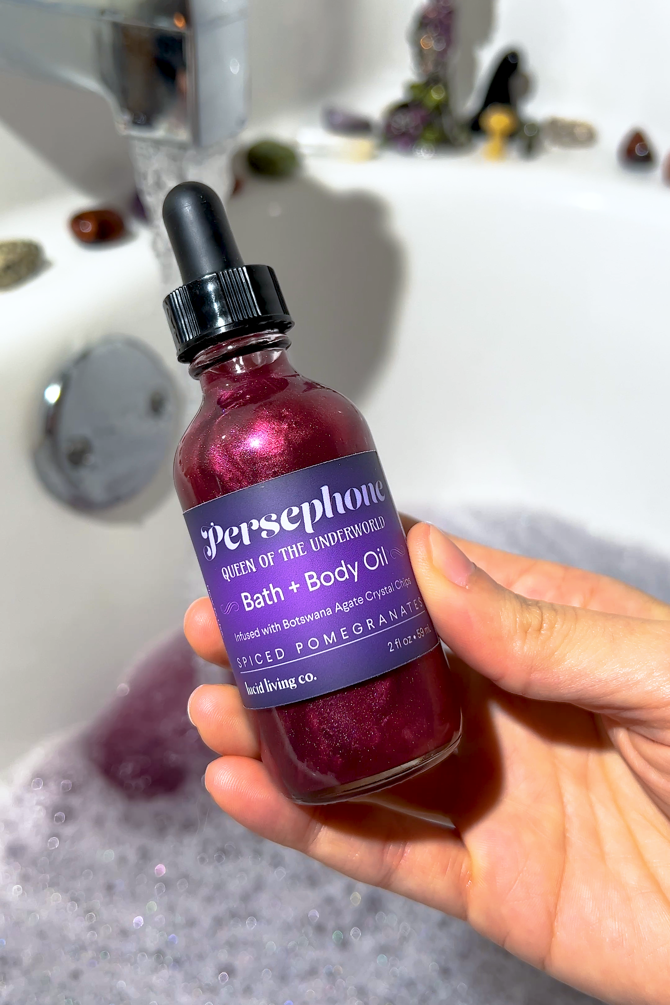 Bottle of Persephone Bath & Body Oil with spiced pomegranate and Botswana Agate crystal chips held over a bubble bath.
