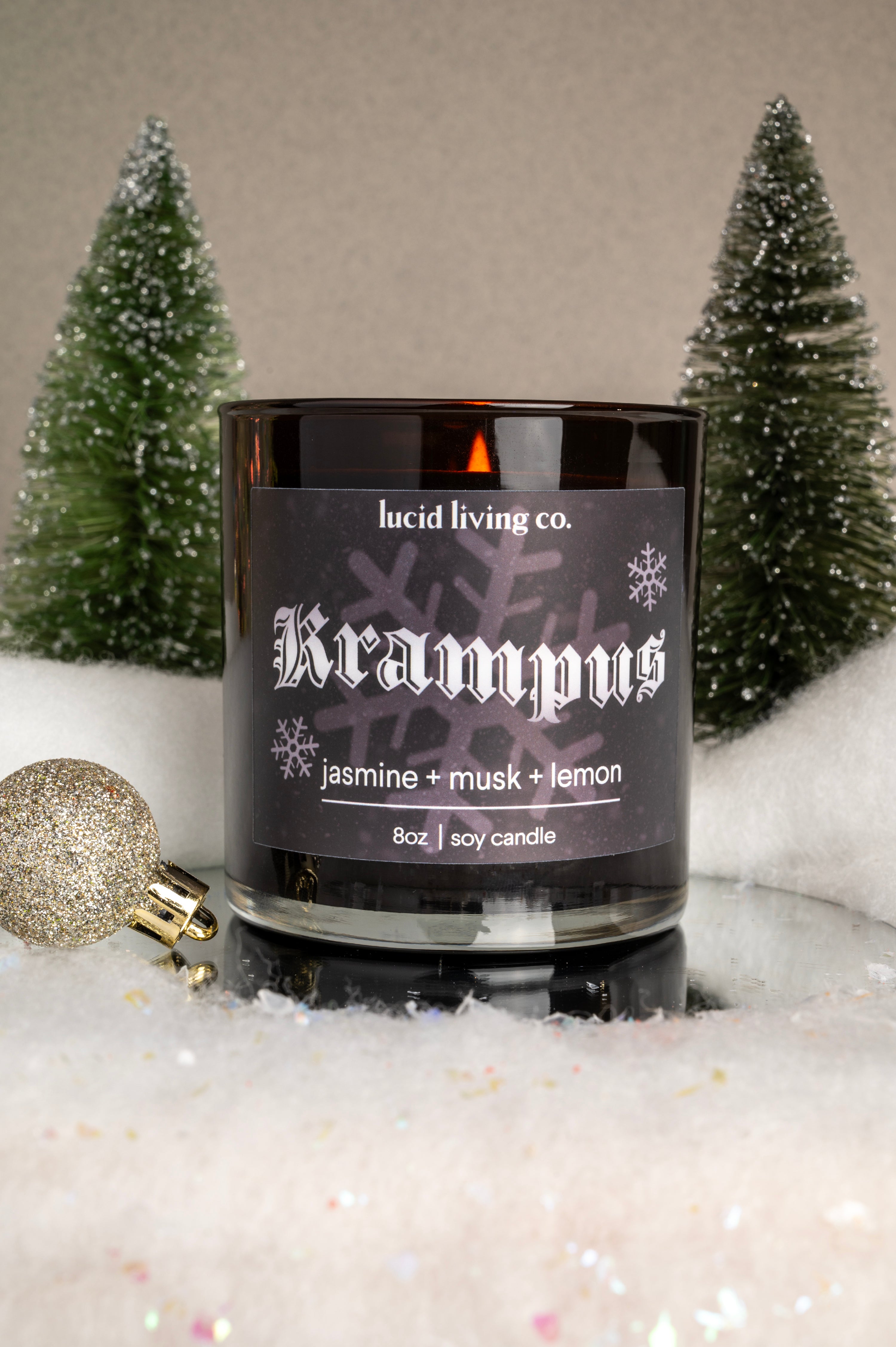 Krampus Soy Candle with jasmine musk and lemon scent in black jar surrounded by snow and holiday decorations