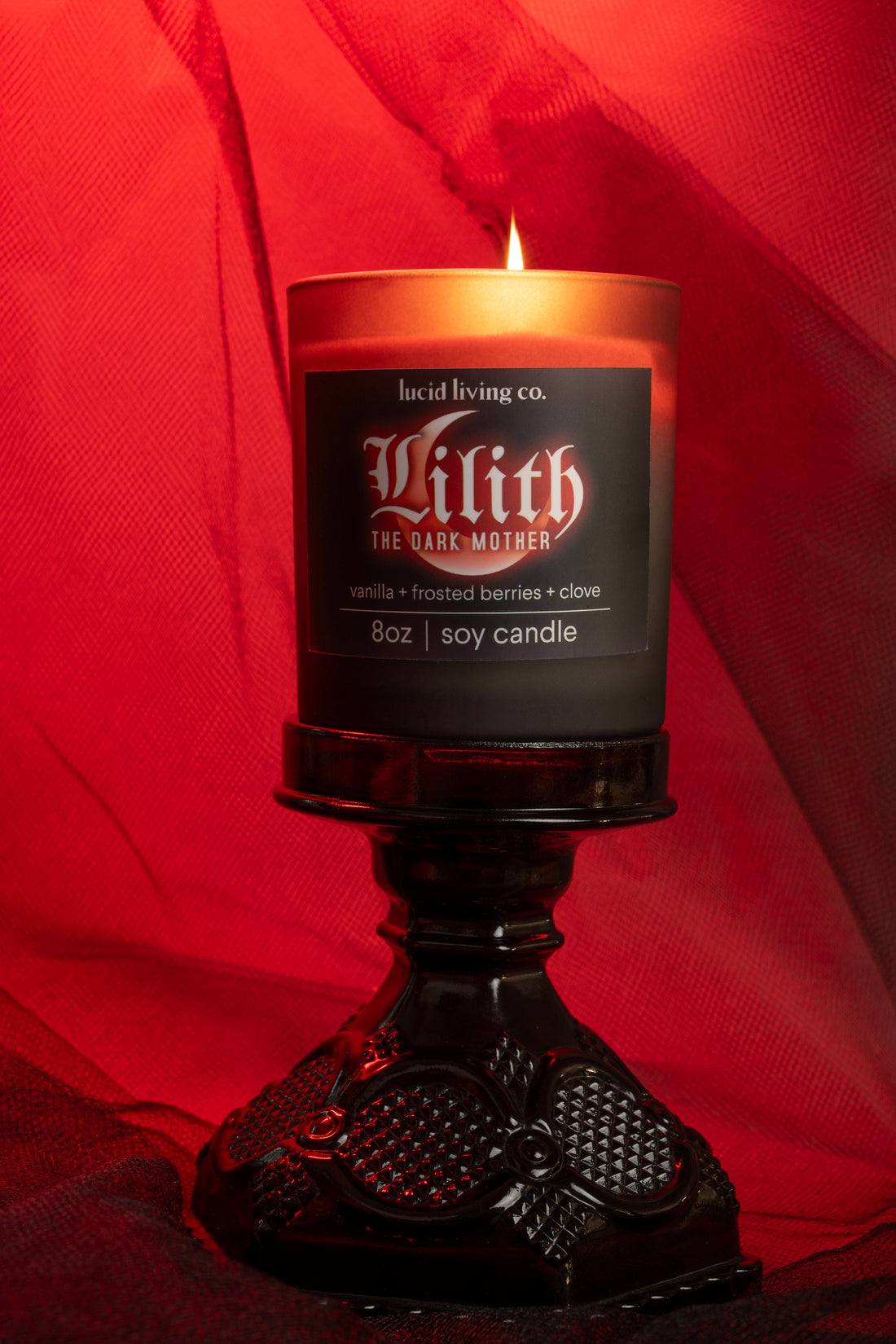 Lilith Soy Candle with vanilla, frosted berries, and clove scent glowing on ornate black stand against red backdrop