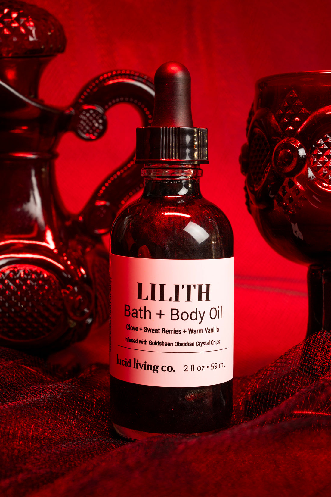 LILITH Bath & Body Oil bottle with dropper surrounded by red decorative glassware on red fabric background.
