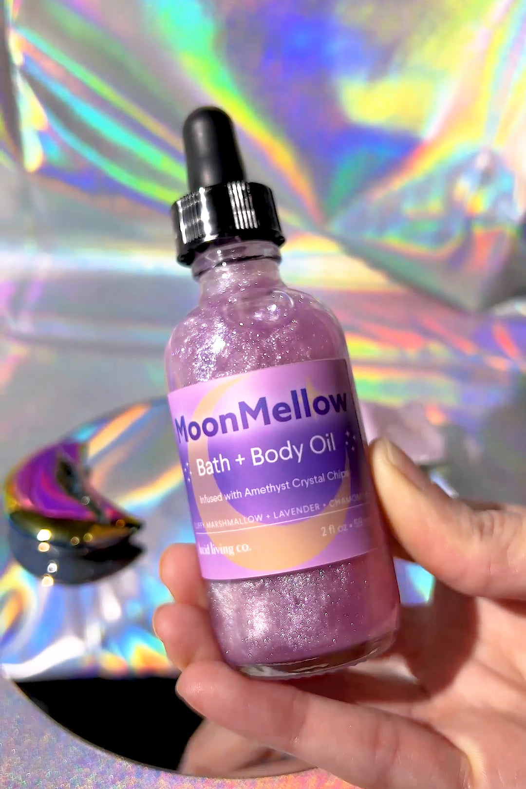 Hand holding MoonMellow Bath & Body Oil bottle with amethyst crystal chips and shimmering purple liquid inside.