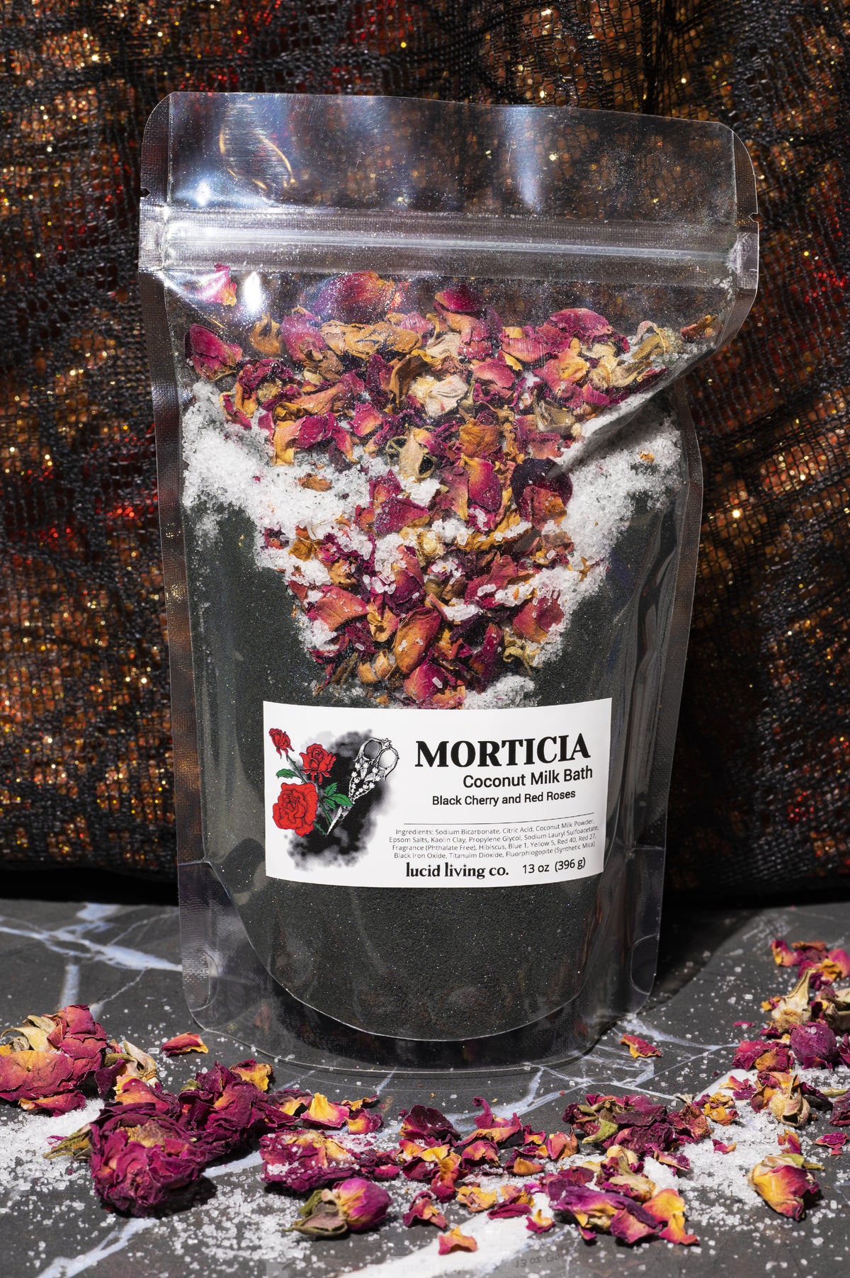 Clear resealable pouch of Morticia Bath Potion featuring black coconut milk bath powder topped with dried rose petals and scattered petals on marble surface with dark fabric background.