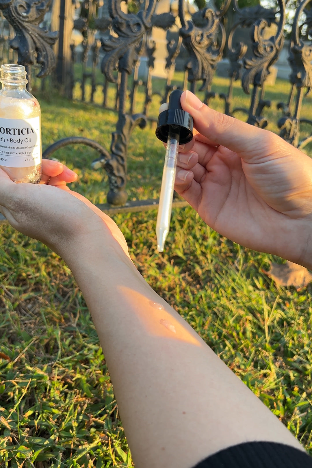 MORTICIA Bath & Body Oil being applied with a dropper onto the forearm outdoors in natural sunlight, highlighting its clear, soothing texture.