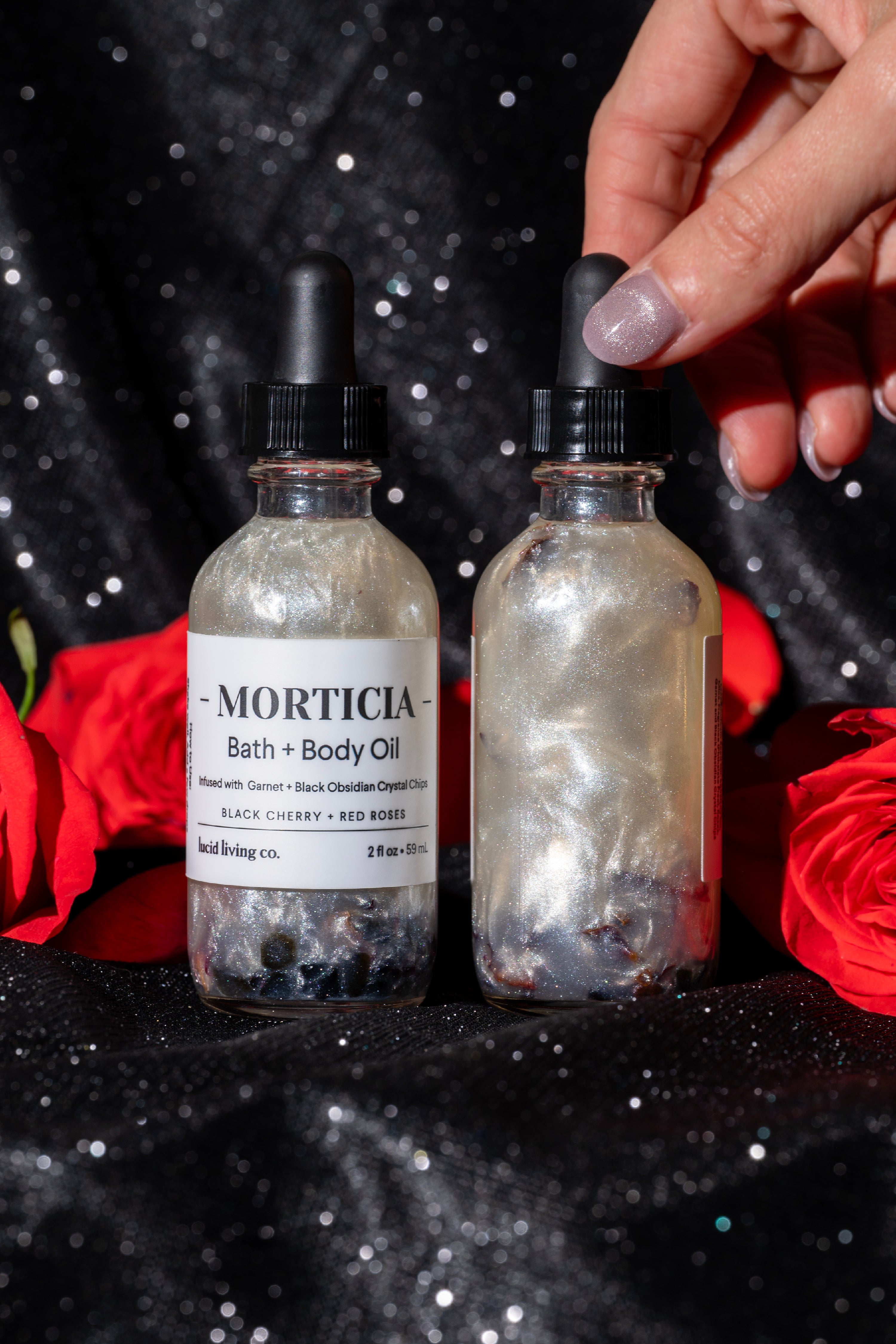Two glass bottles of MORTICIA Bath & Body Oil with shimmering liquid and black obsidian chips inside, set against a black sparkling fabric with red roses and a hand adjusting a dropper cap.