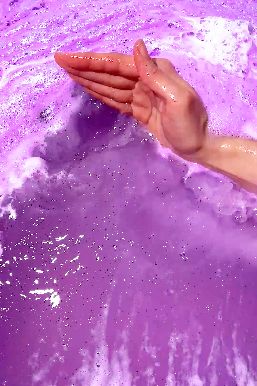 Mystical Fae Bath Potion creating vibrant purple bubbles as hand stirs soothing honey and lilac blossom scented water.