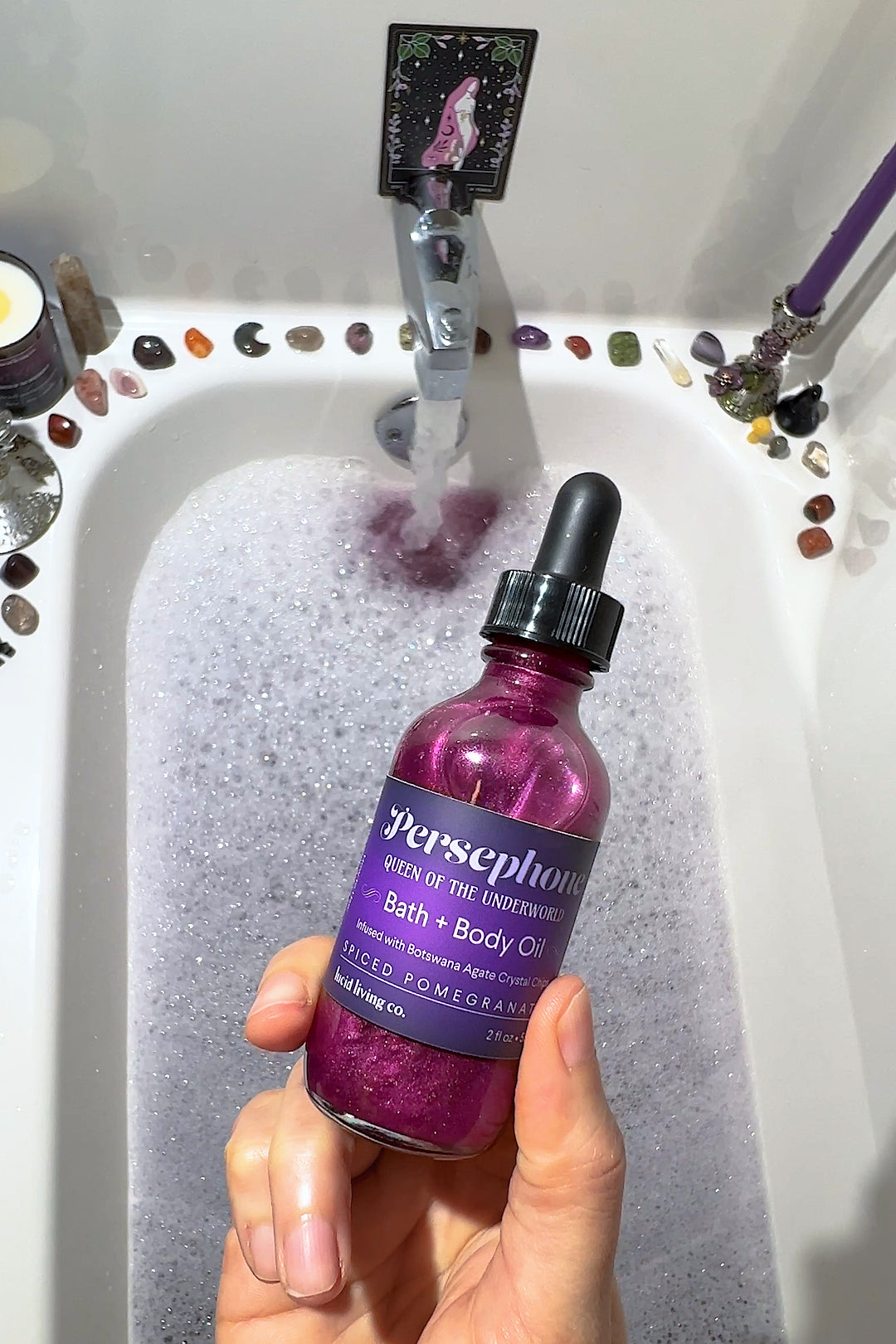 Hand holding Persephone Bath & Body Oil bottle over a foamy bathtub with crystals and candles around.