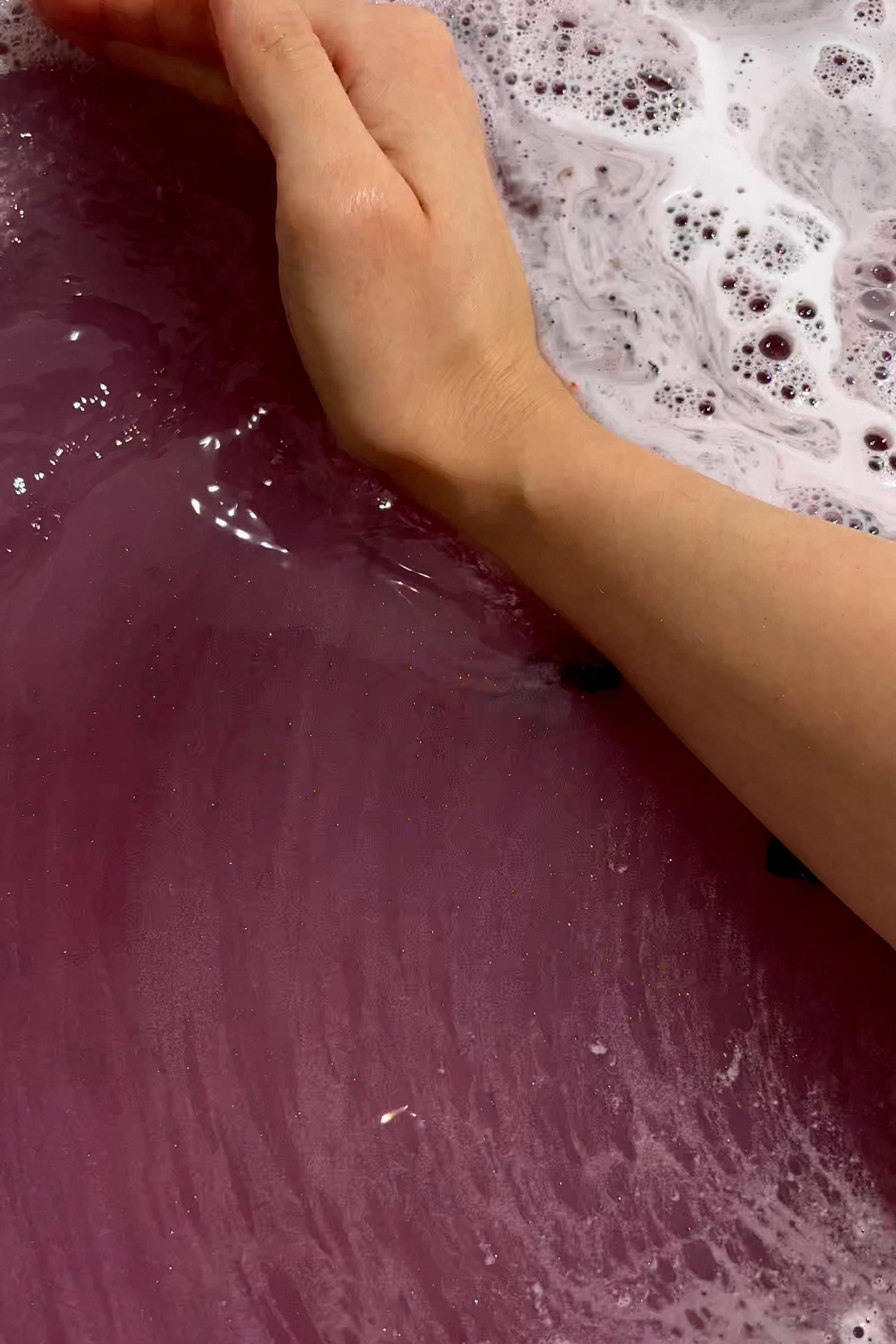 Hand touching purple water with foam from the PERSEPHONE "Queen of the Underworld" Coconut Milk Bath product.