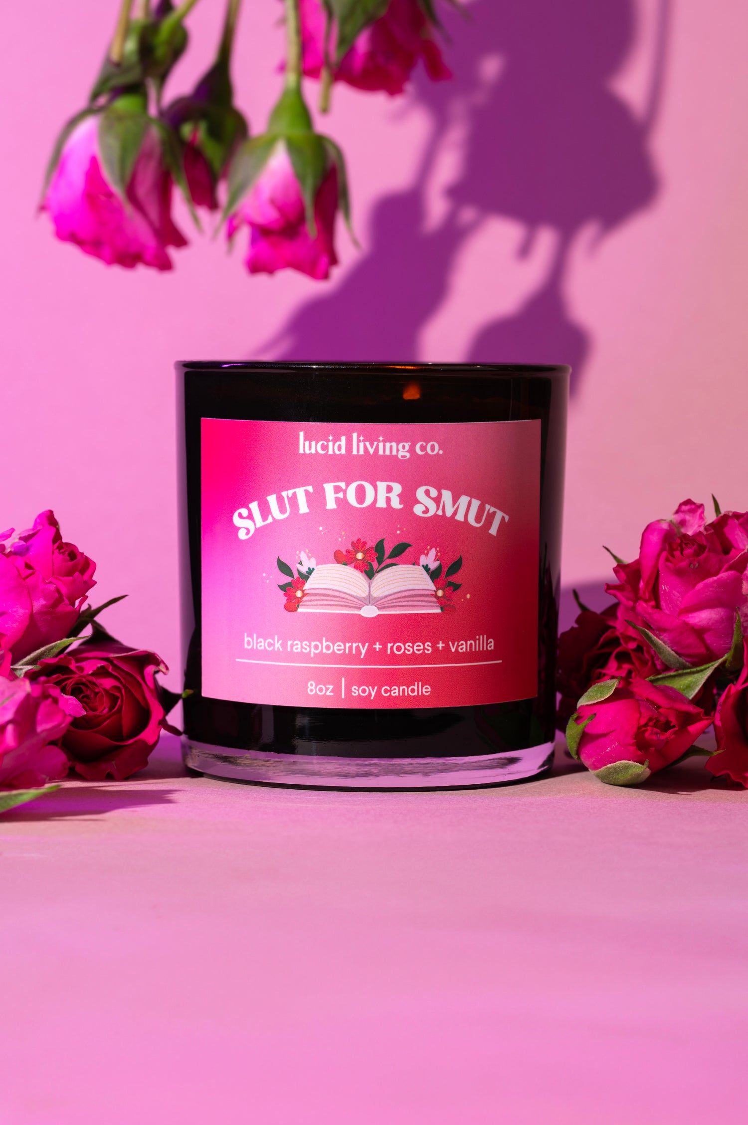 Slut for Smut Soy Candle with black raspberry, roses, and vanilla scent surrounded by pink roses on pink background.