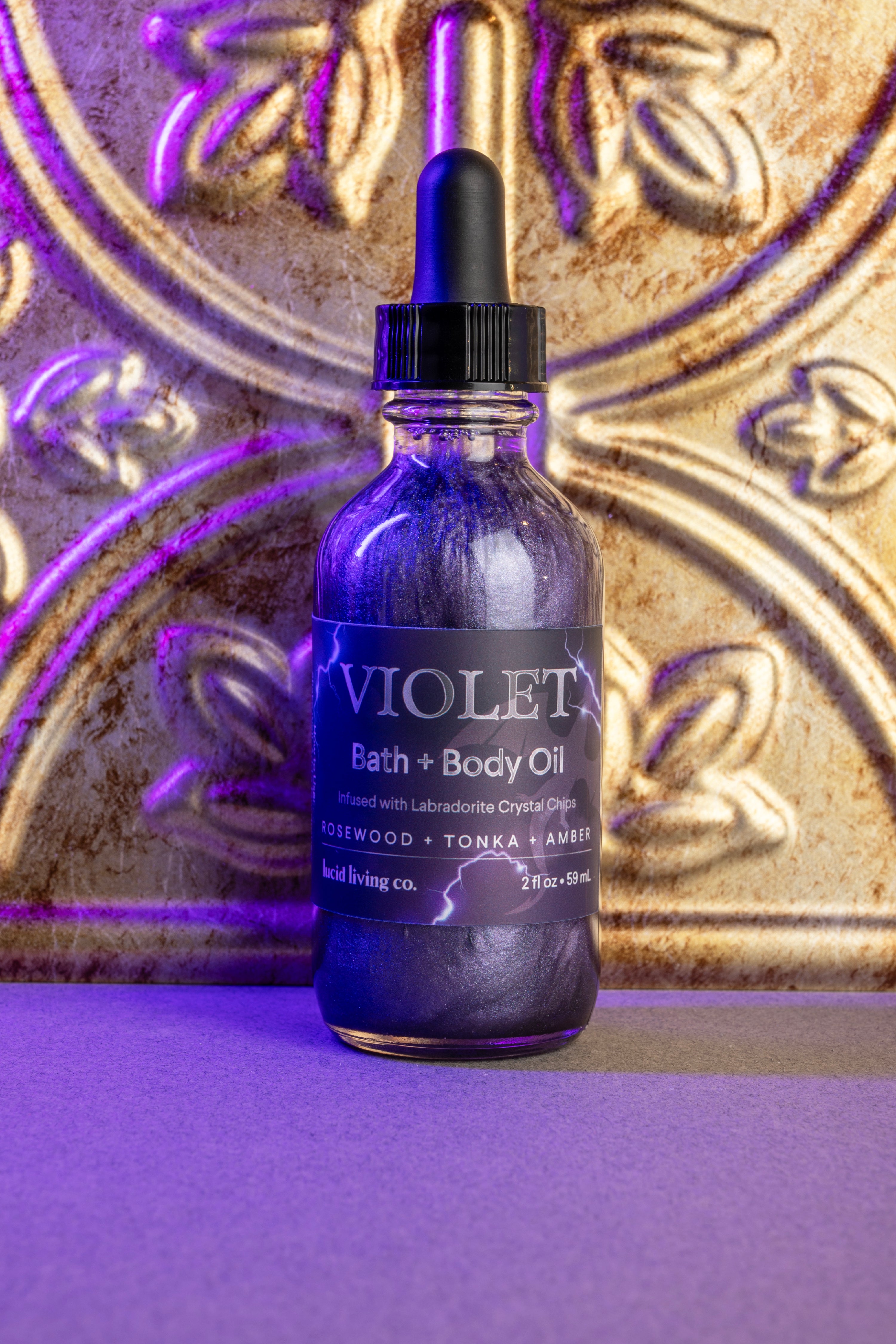 Violet Bath & Body Oil bottle with shimmering liquid and dark amber and violet lighting on textured background.