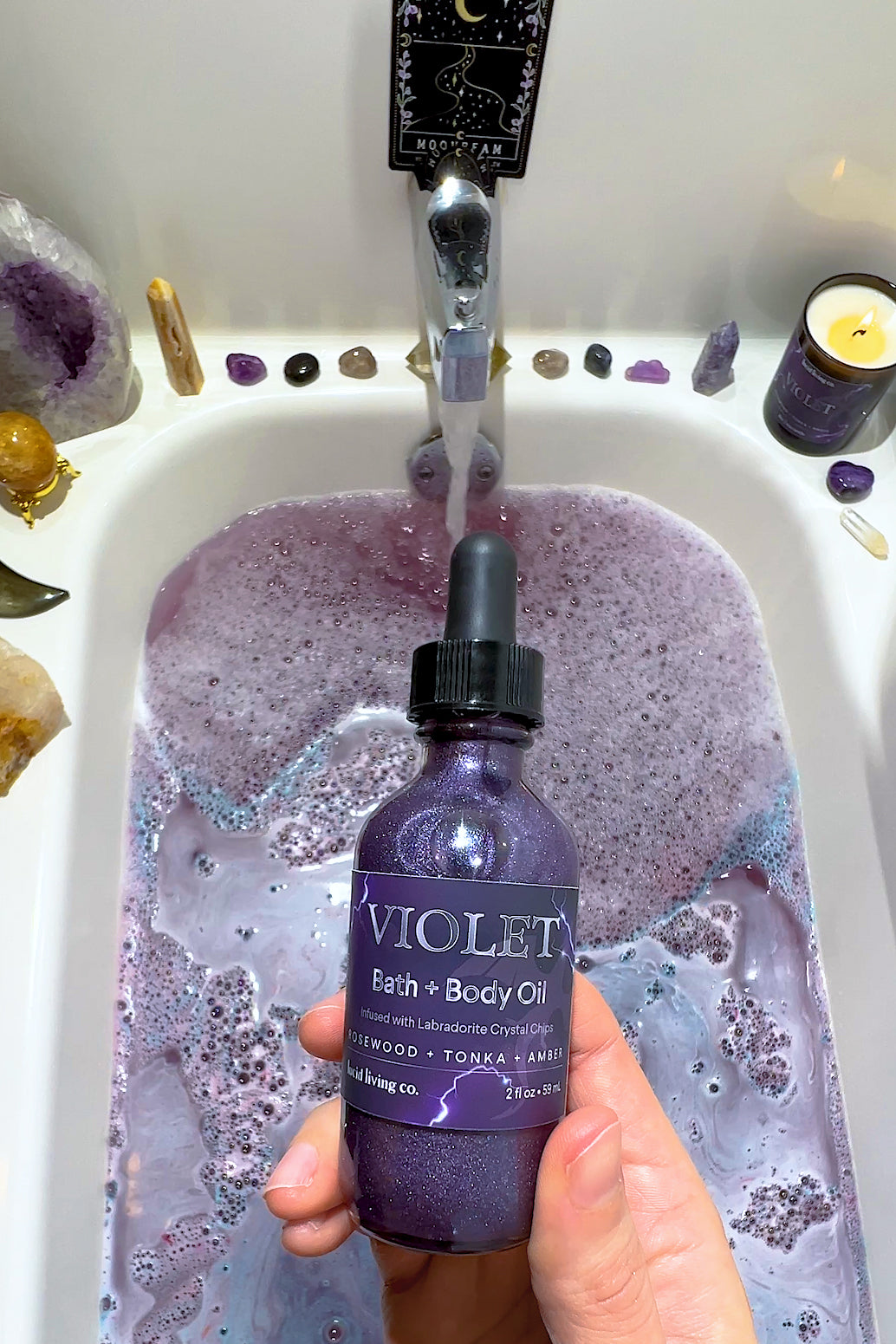 Violet Bath & Body Oil bottle held over purple foamy bath surrounded by crystals and lit candle.