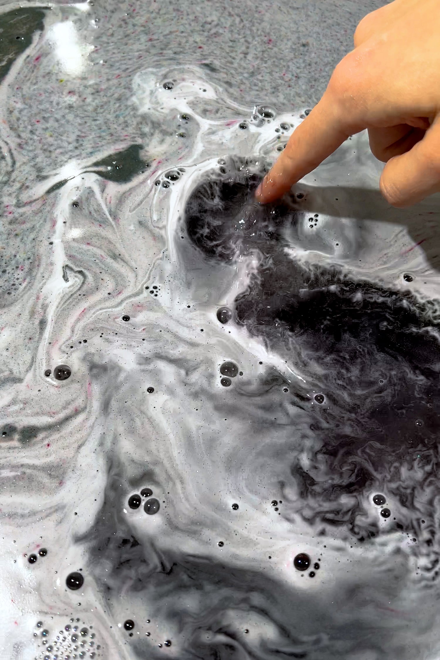 Finger touching swirling bubbles and colors in bathwater with LILITH "The Dark Mother" Bath Potion dissolving.