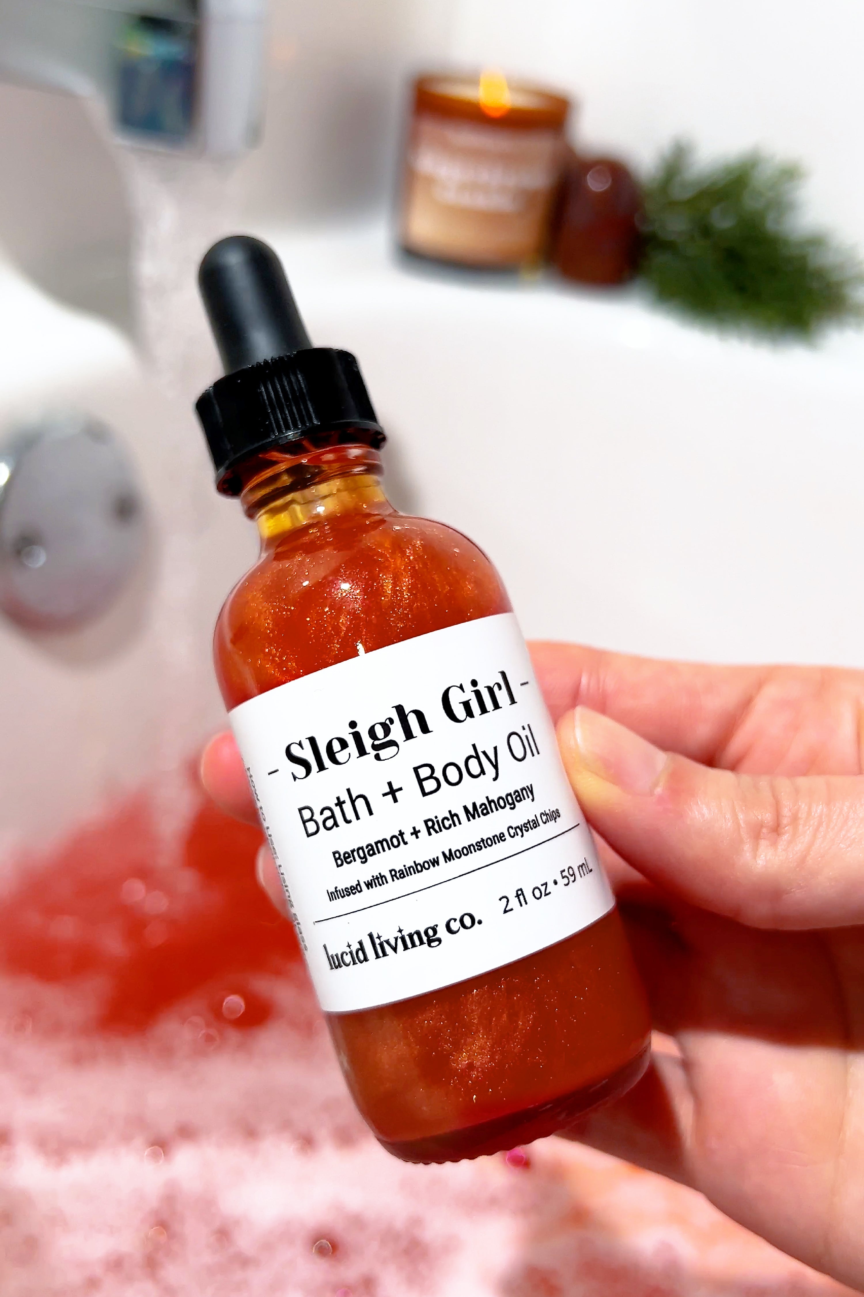 Hand holding Sleigh Girl Bath & Body Oil bottle with shimmery glow in a pink bath setting.