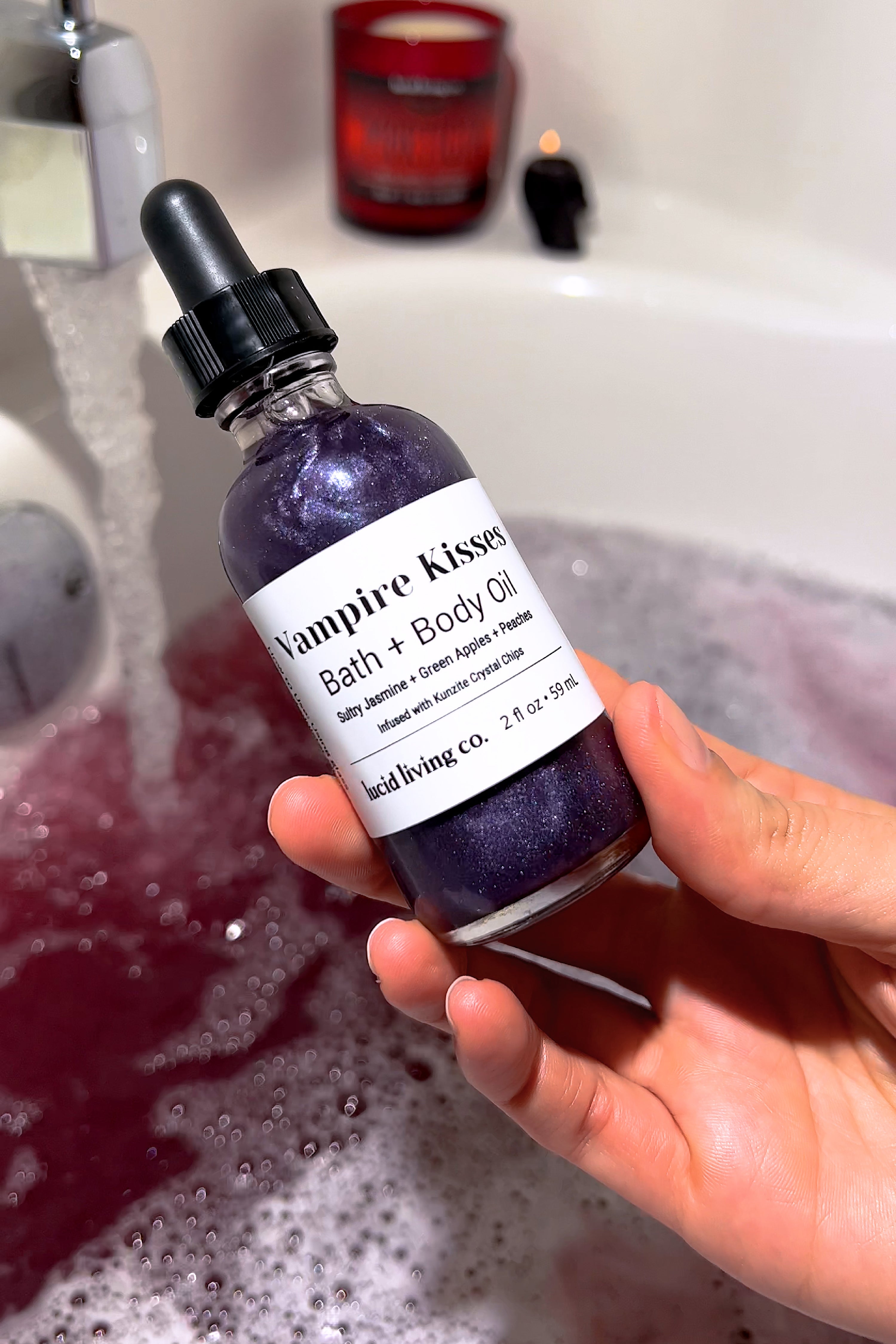 Bottle of Vampire Kisses Bath & Body Oil with shimmering purple liquid held over a pink bubble bath.