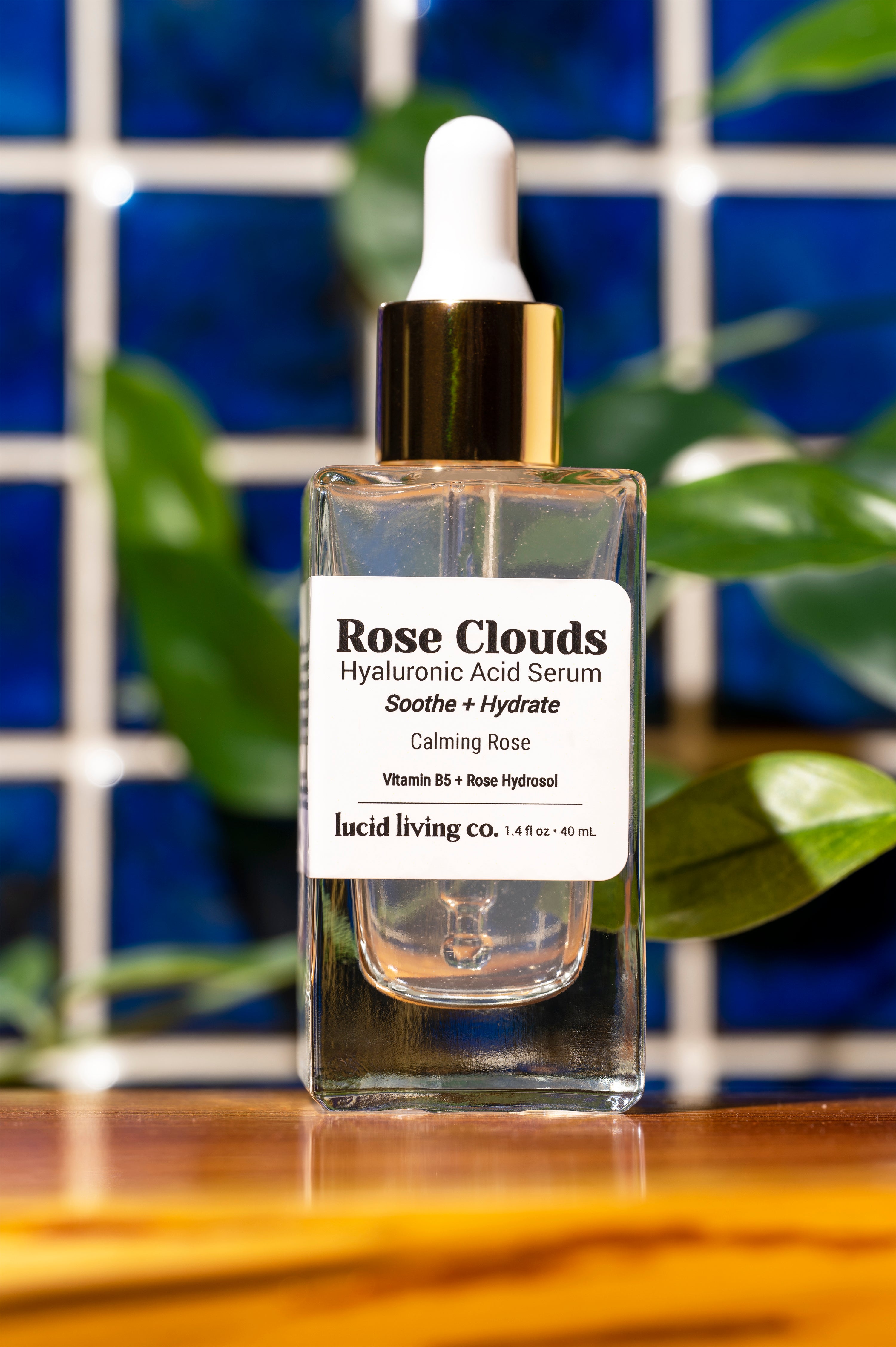 Rose Clouds Hyaluronic Acid Serum in clear glass bottle with dropper and gold collar on wooden surface with plants background