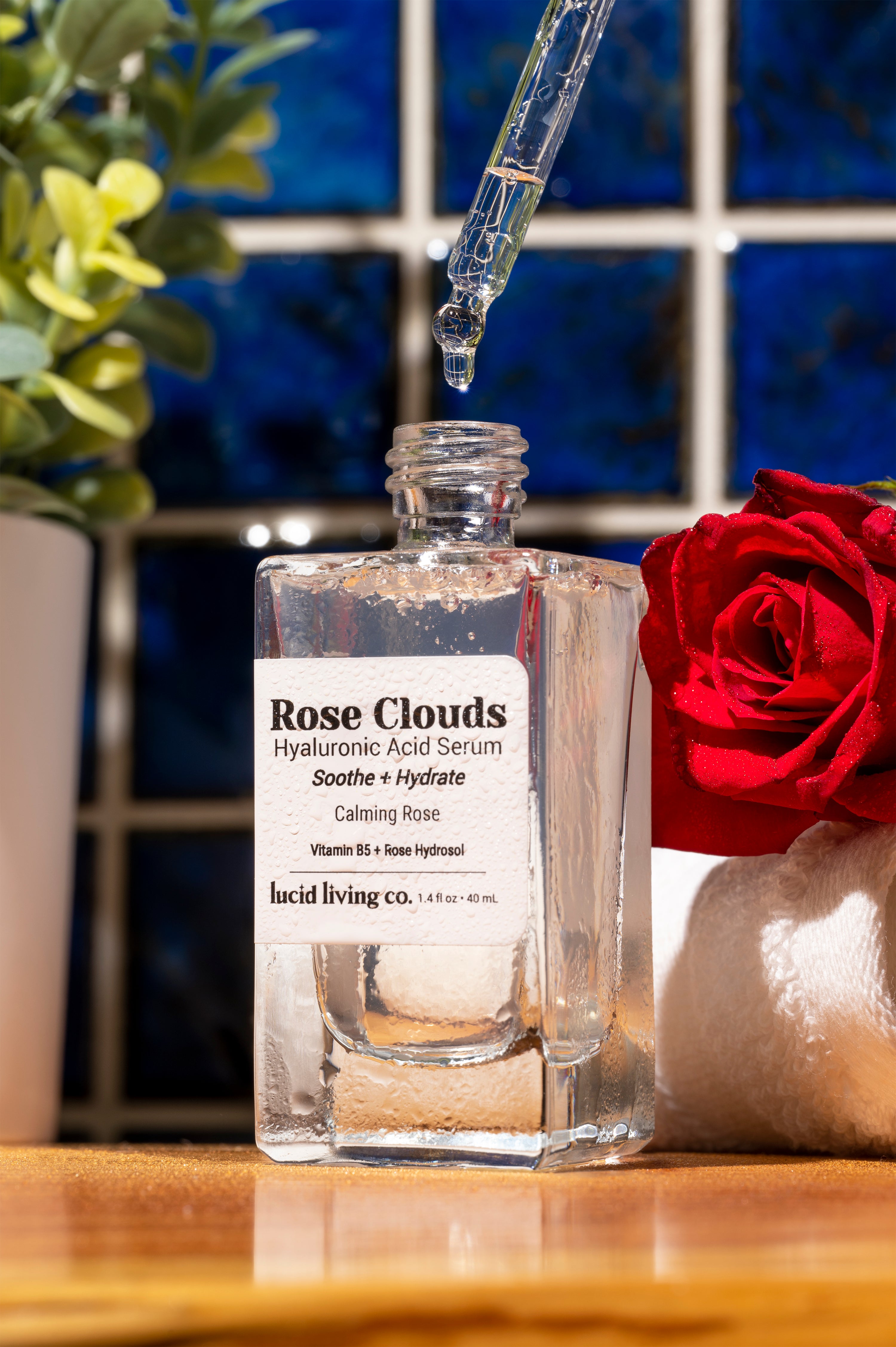 Rose Clouds Hyaluronic Acid Serum bottle with dropper releasing serum, next to a red rose on a wooden surface.