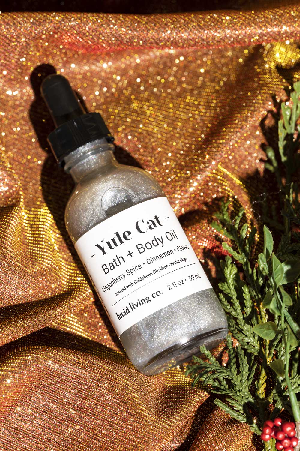 Yule Cat Bath & Body Oil bottle on glittery fabric with greenery and red berries beside it.