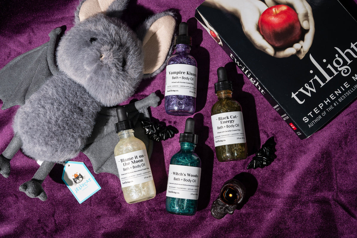 Two bottles of 'Pumpkin Cat' and 'Black Cat Energy' bath & body oils on a wooden board with a cat and candle in the background.