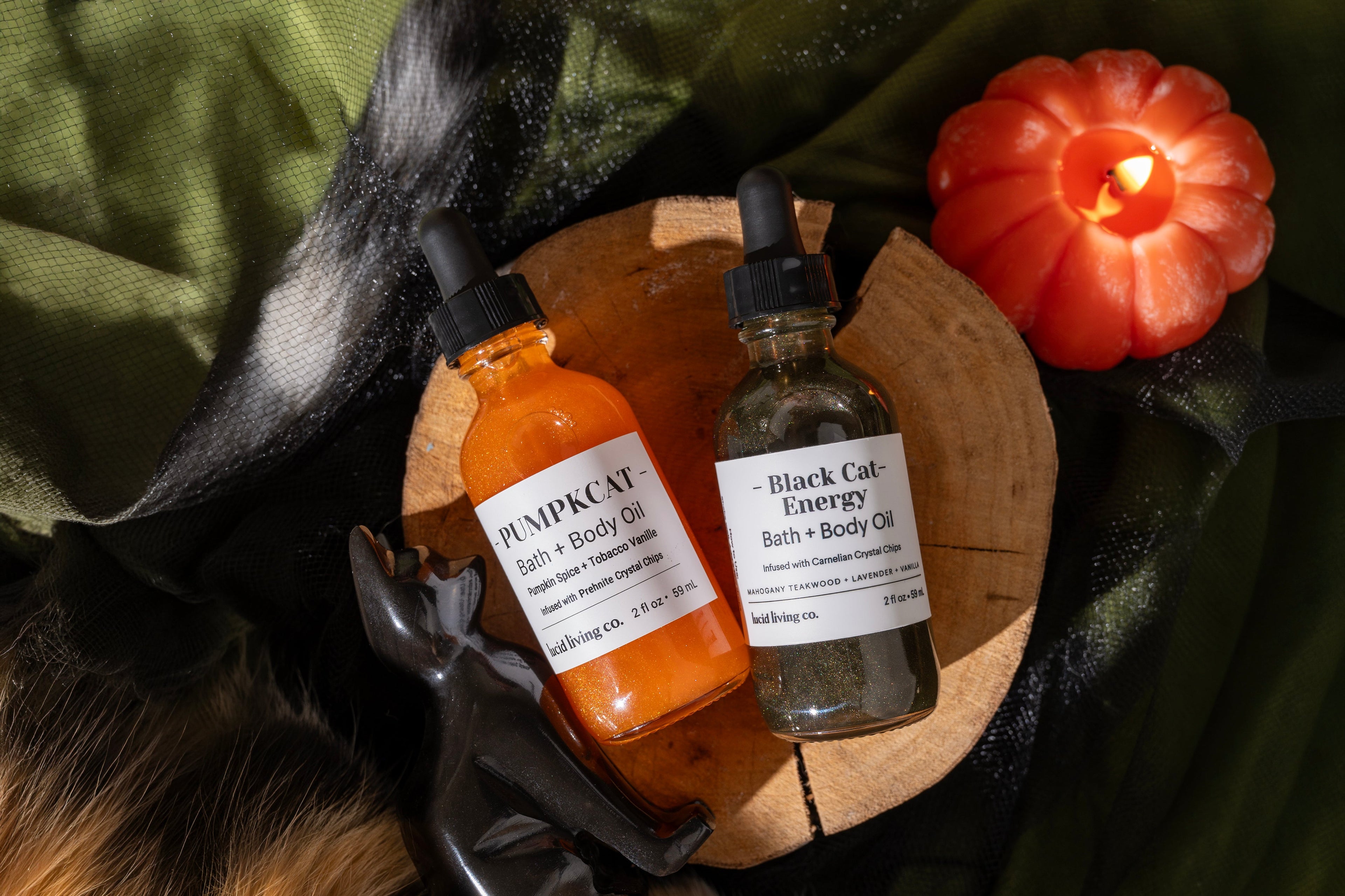 Two bottles of 'Pumpkin Cat' and 'Black Cat Energy' bath & body oils on a wooden board with a cat and candle in the background.