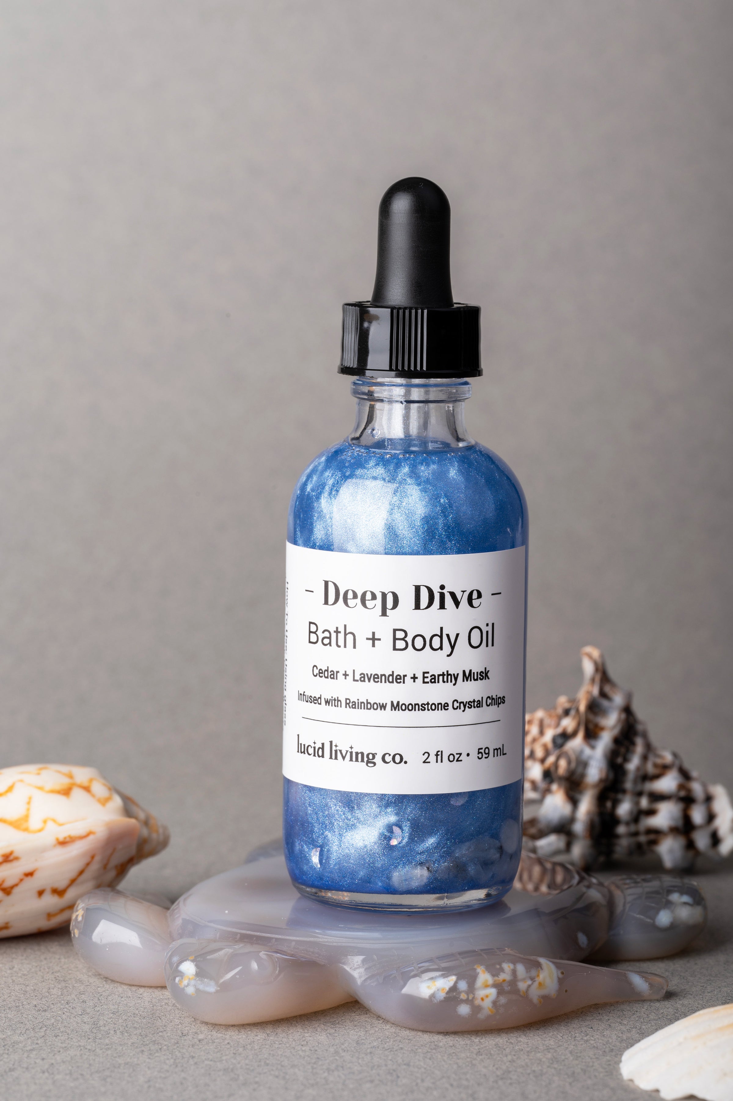 Clear glass bottle of Deep Dive Bath + Body Oil with shimmering blue liquid and black dropper, accented with seashells and stones, showcasing the Midnight Depths Bundle.