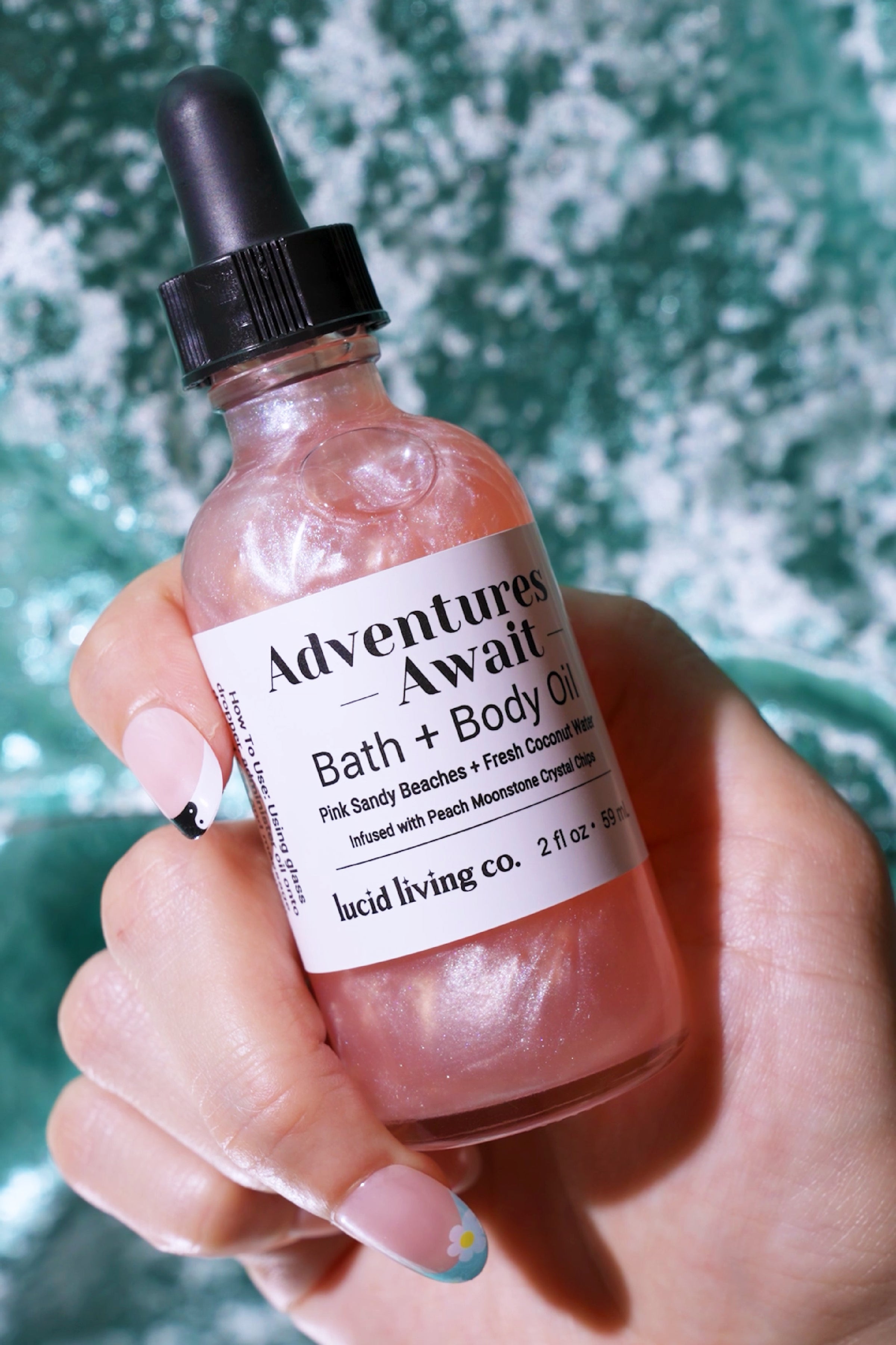 Hand holding a pearlescent pink bottle of Adventures Await Bath & Body Oil with a black dropper cap, shimmering against a textured green and white background.