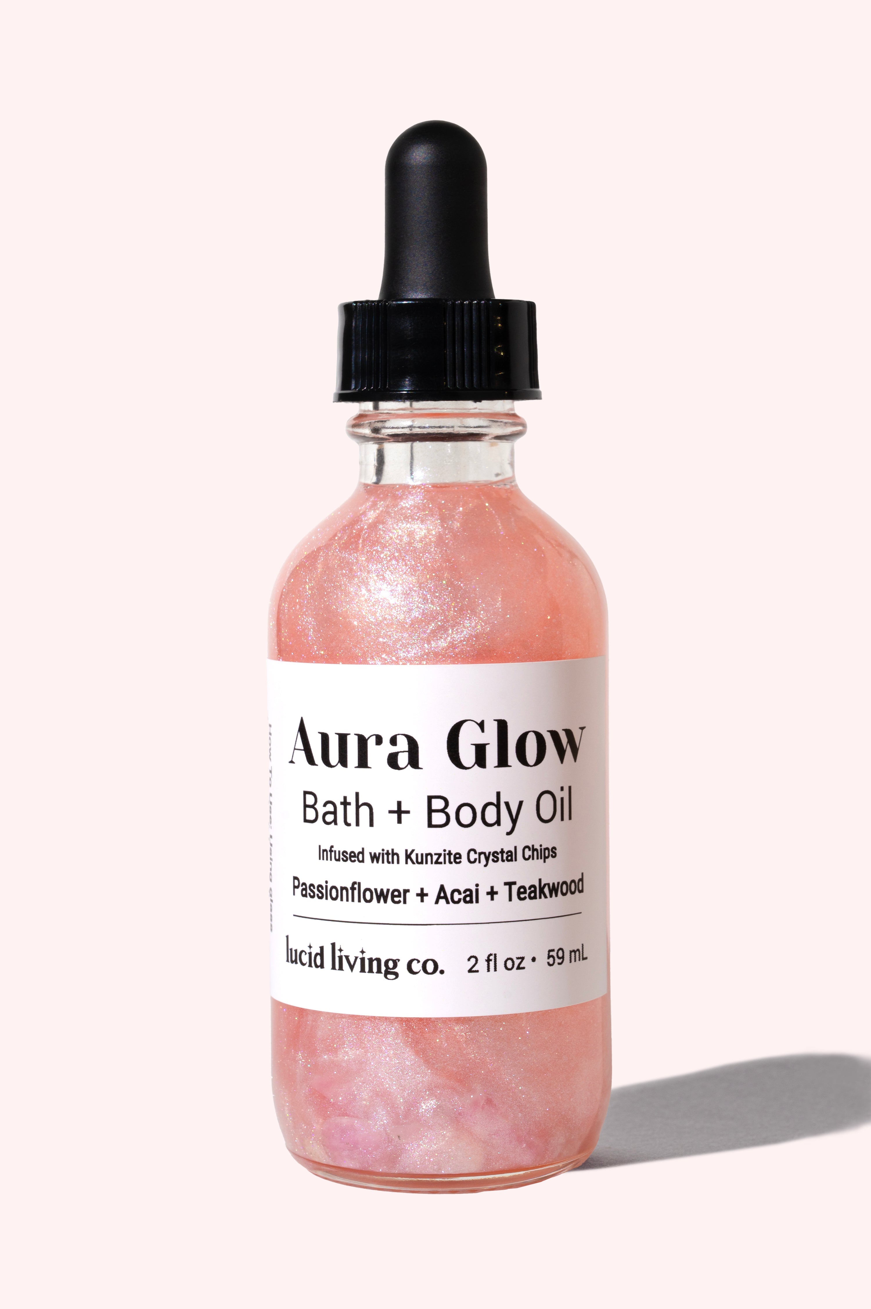 Clear glass bottle of Aura Glow Bath &amp; Body Oil with shimmering pink liquid and a black dropper cap, set against a soft pale background, highlighting its luminous glow.