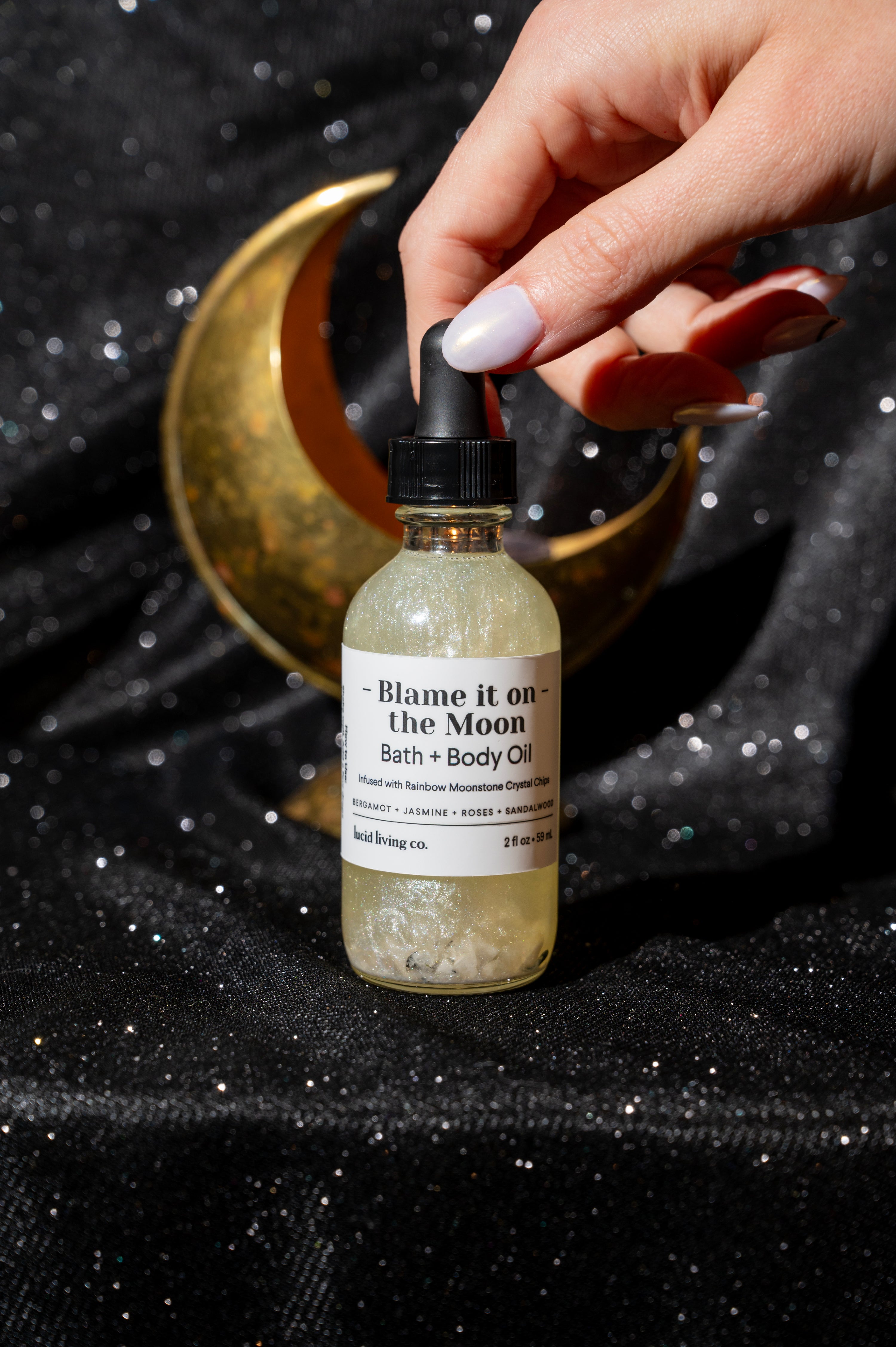 Hand holding a shimmering Blame it on the Moon Bath &amp; Body Oil bottle with a black dropper cap, set against a sparkling black fabric backdrop featuring a decorative gold crescent moon.