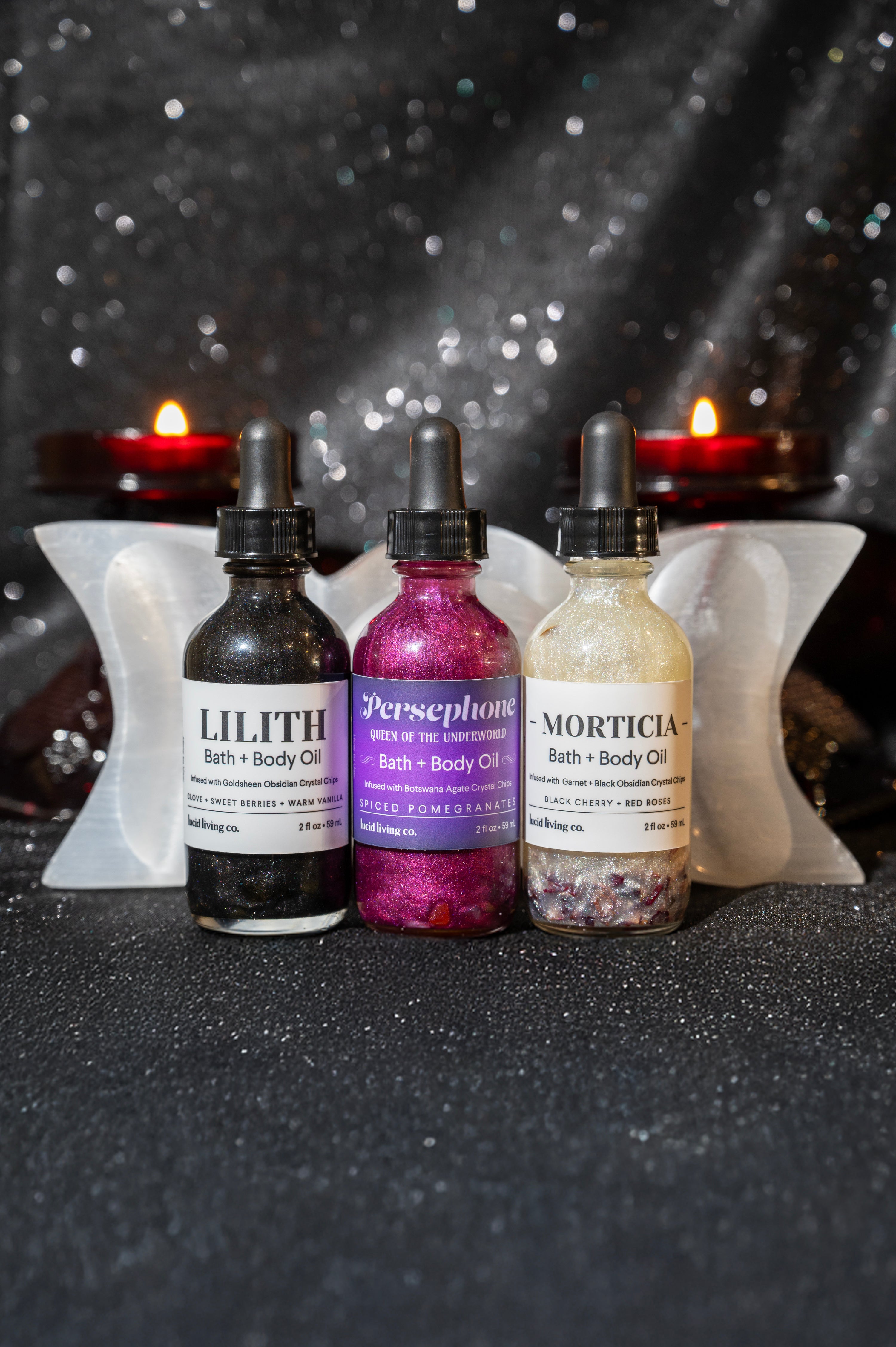Three shimmering bottles of body oil named Lilith, Persephone, and Morticia arranged on a sparkling dark surface with white angel wings and lit red candles in the background, featuring the Dark Goddess Bundle.