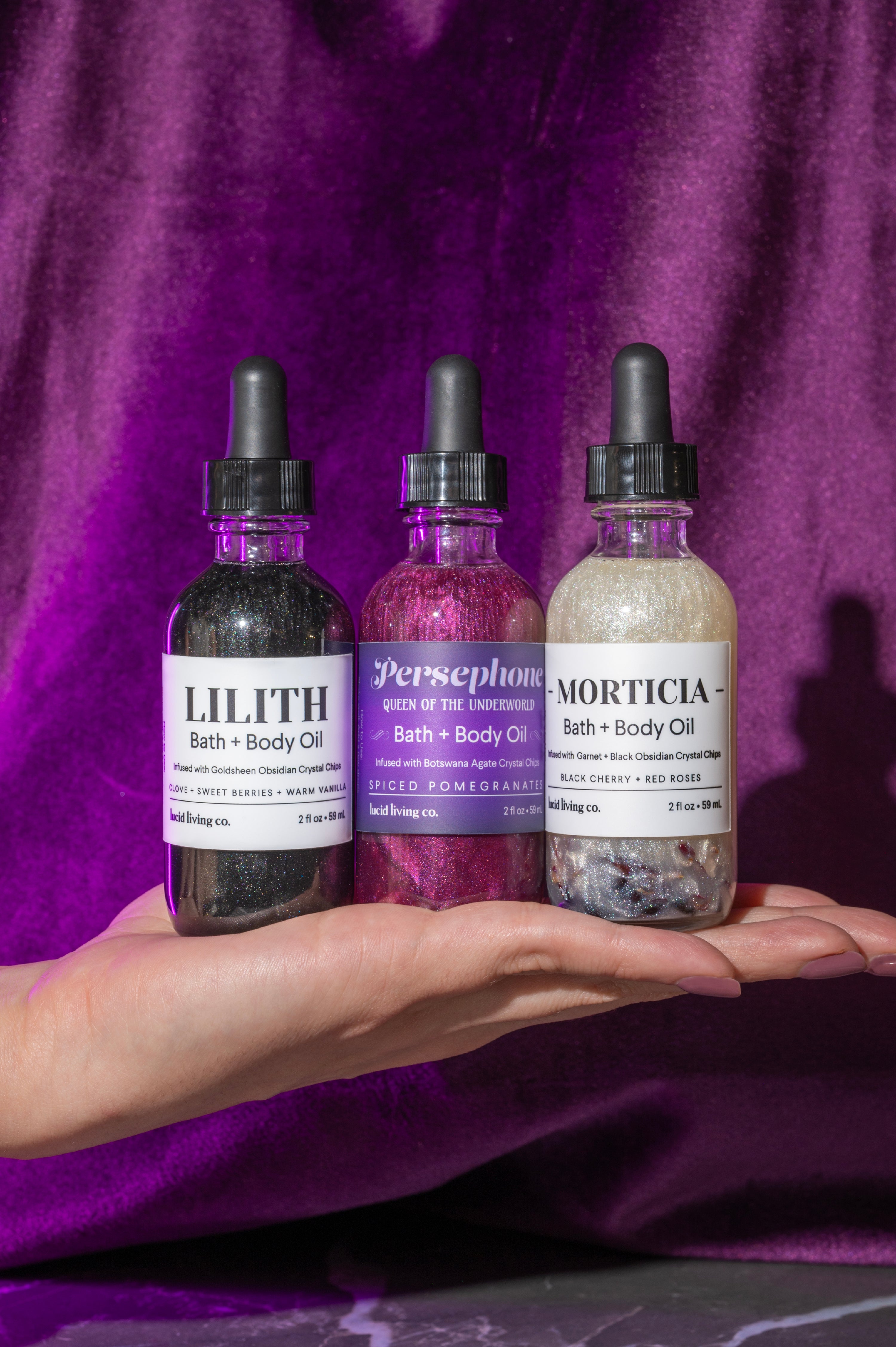 Hand presenting three shimmering bottles of Dark Goddess Bundle Bath + Body Oil, featuring Lilith, Persephone, and Morticia scents, against a rich purple velvet backdrop.