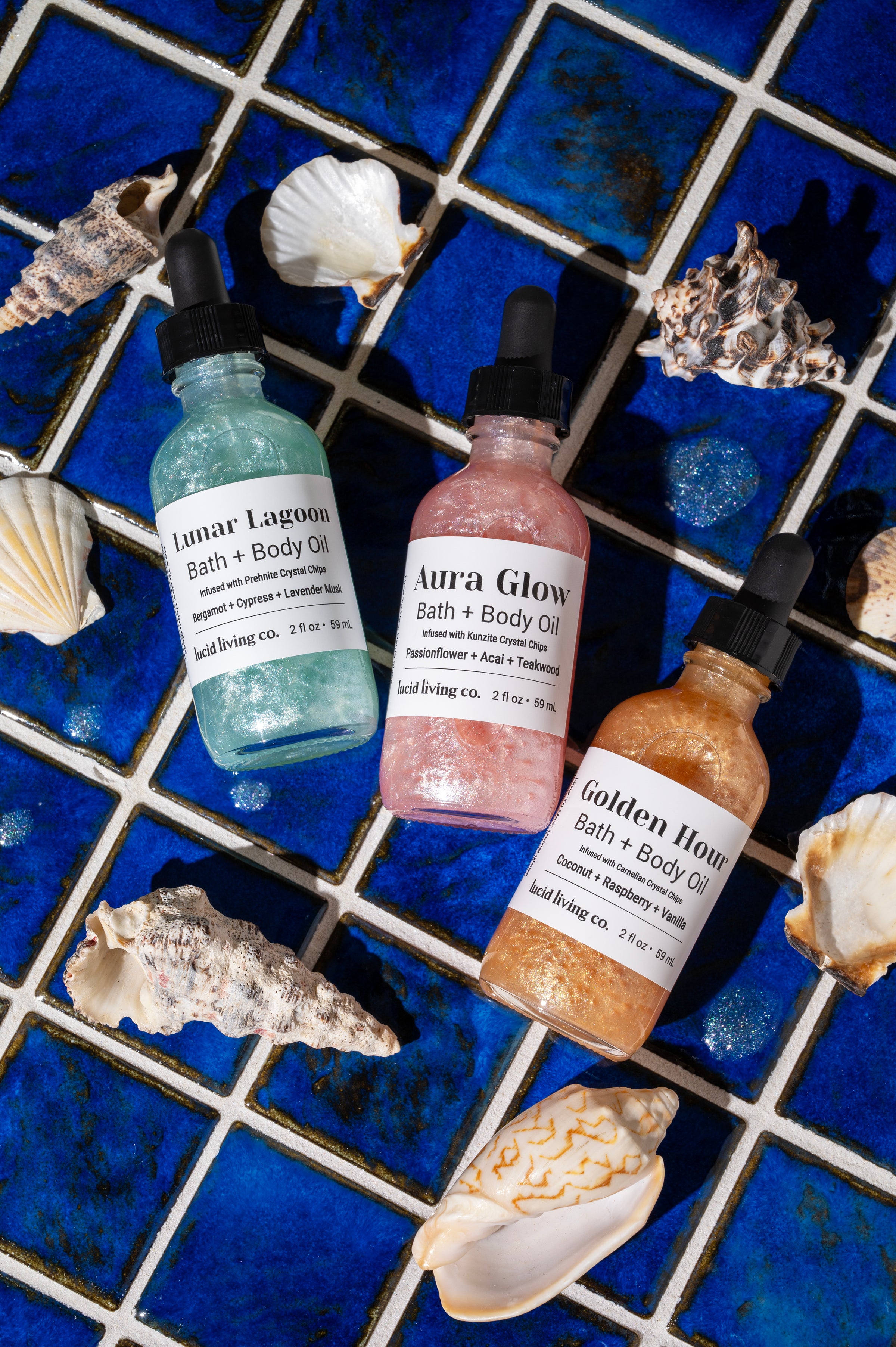 Three shimmering bottles of bath and body oil with crystal chips inside, placed on vibrant blue tiles surrounded by seashells, showcasing the Endless Summer Bundle.
