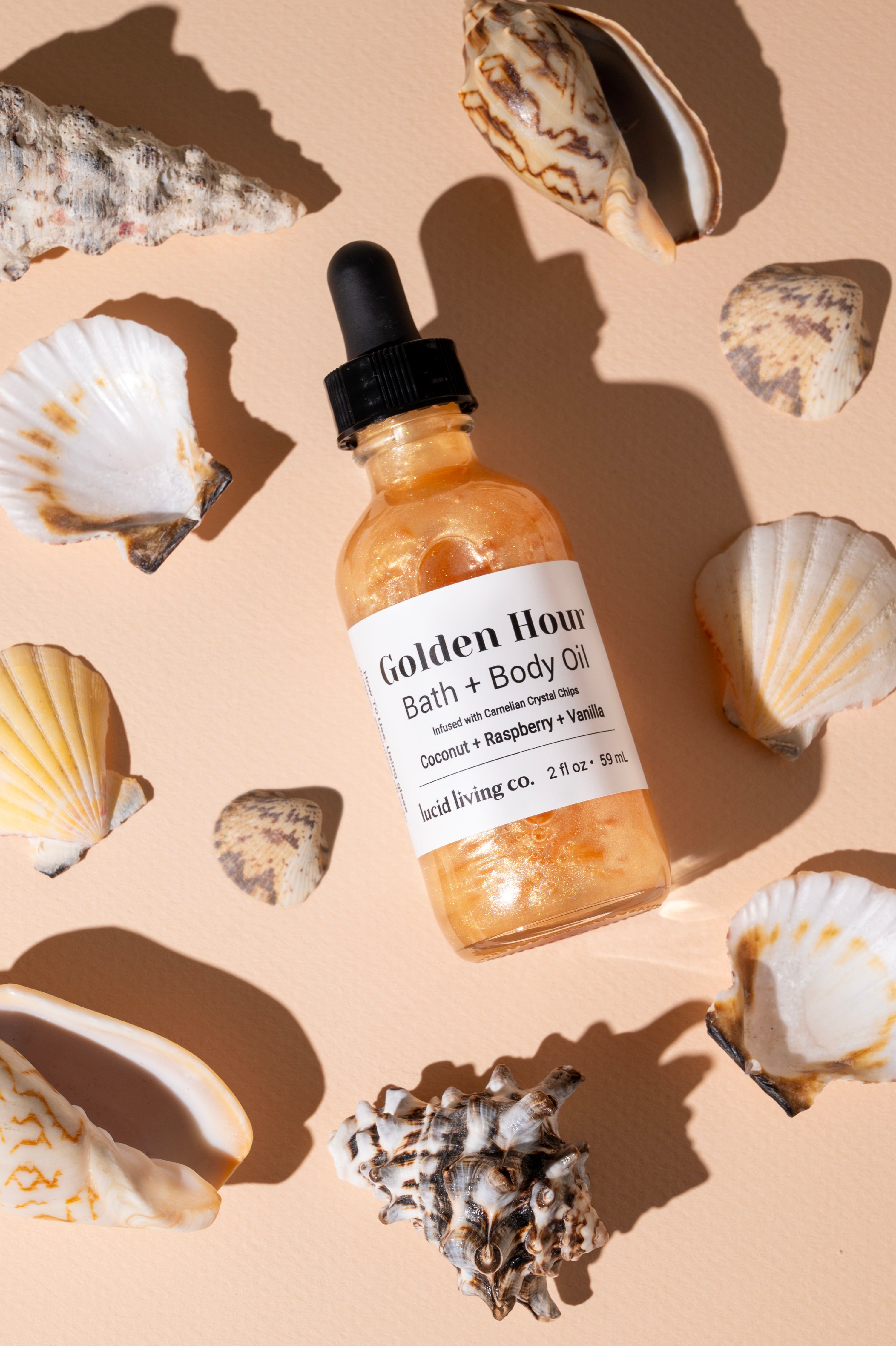 Golden Hour Bath &amp; Body Oil in a clear glass bottle with black dropper, containing shimmering golden liquid, surrounded by various seashells on a warm beige surface.