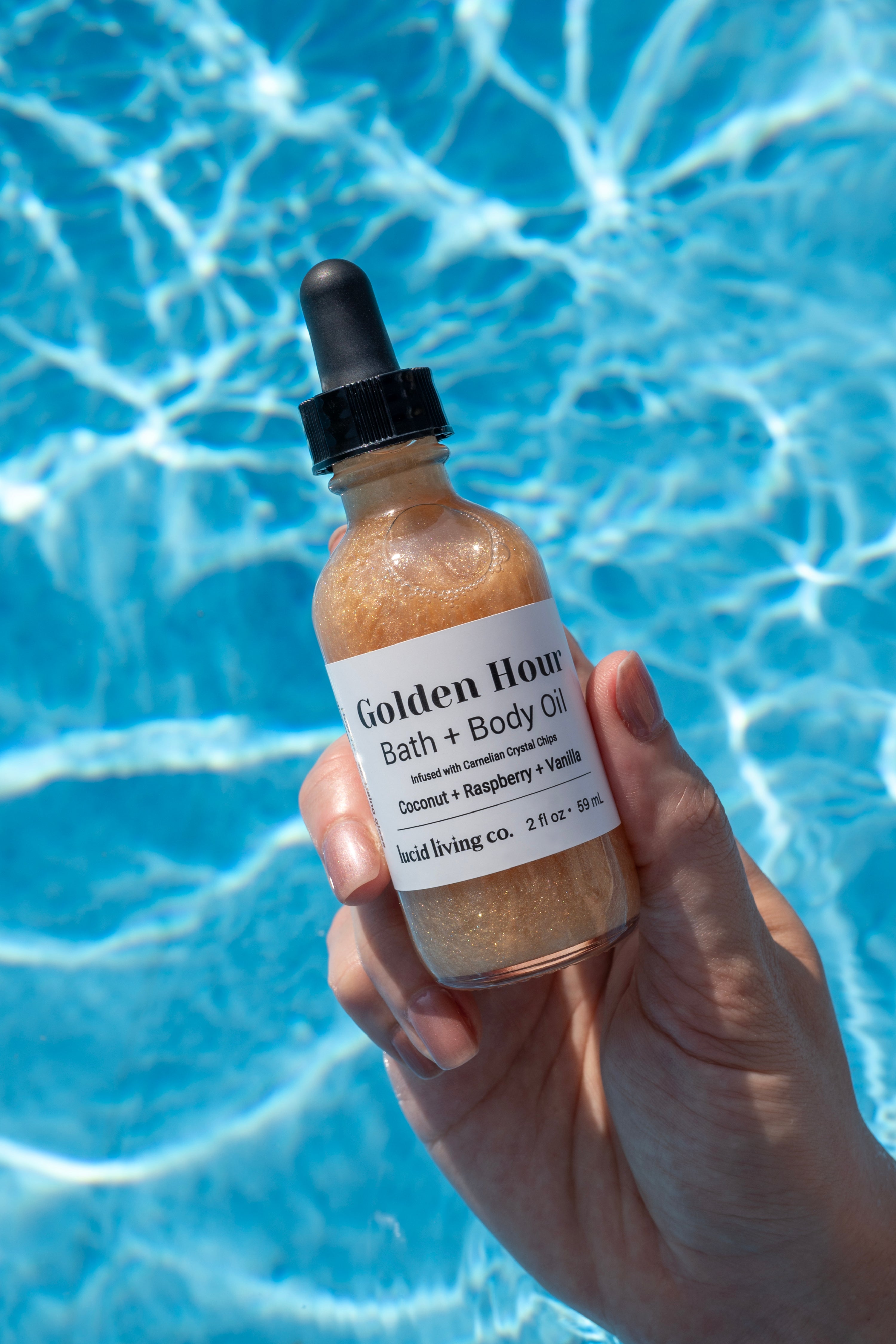 Hand holding a shimmering bottle of Golden Hour Bath &amp; Body Oil with golden liquid and black dropper, set against the bright, rippling blue water of a pool.