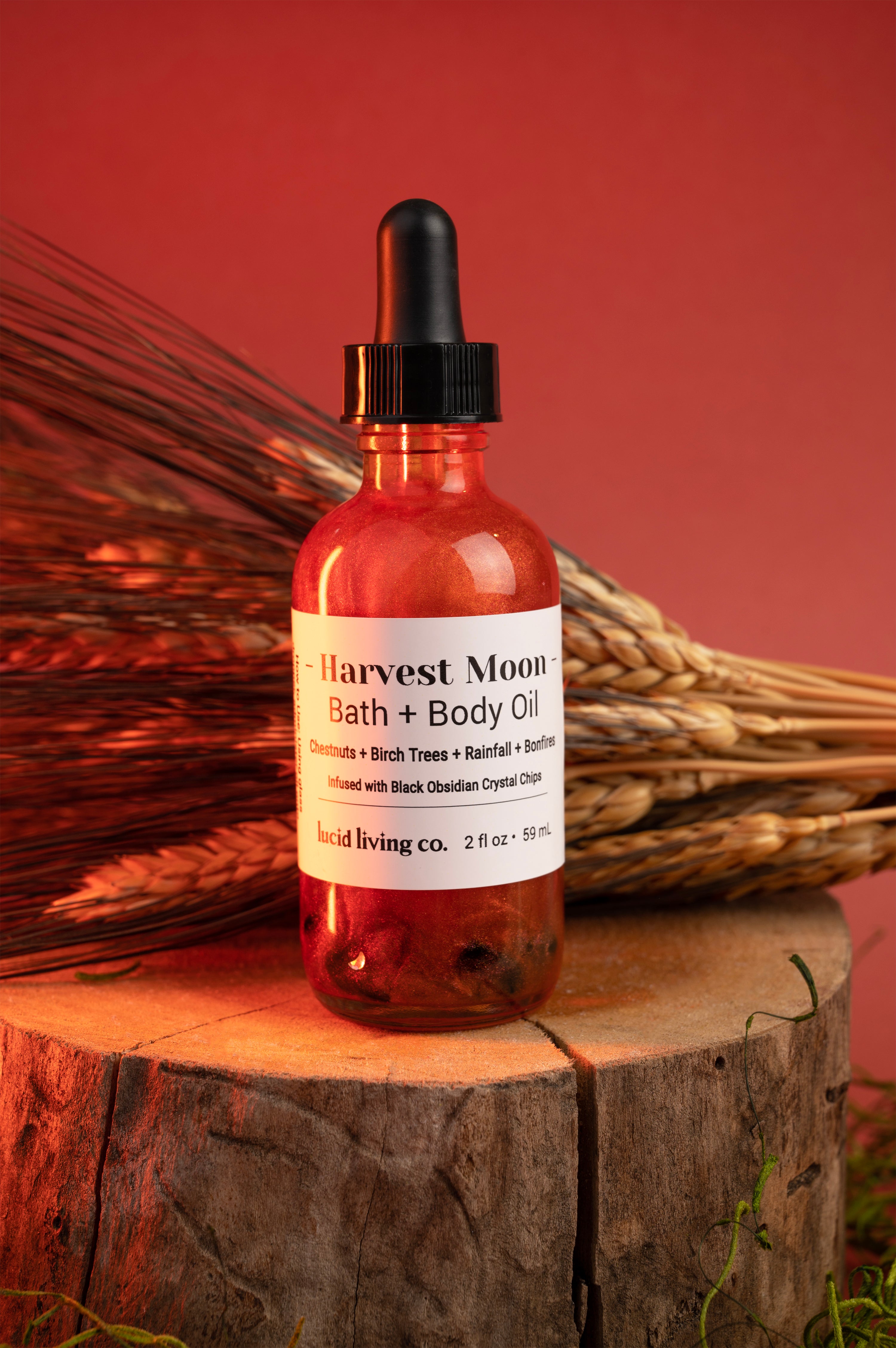 Harvest Moon Body Oil bottle with chestnuts and wheat on wooden stump against warm background.