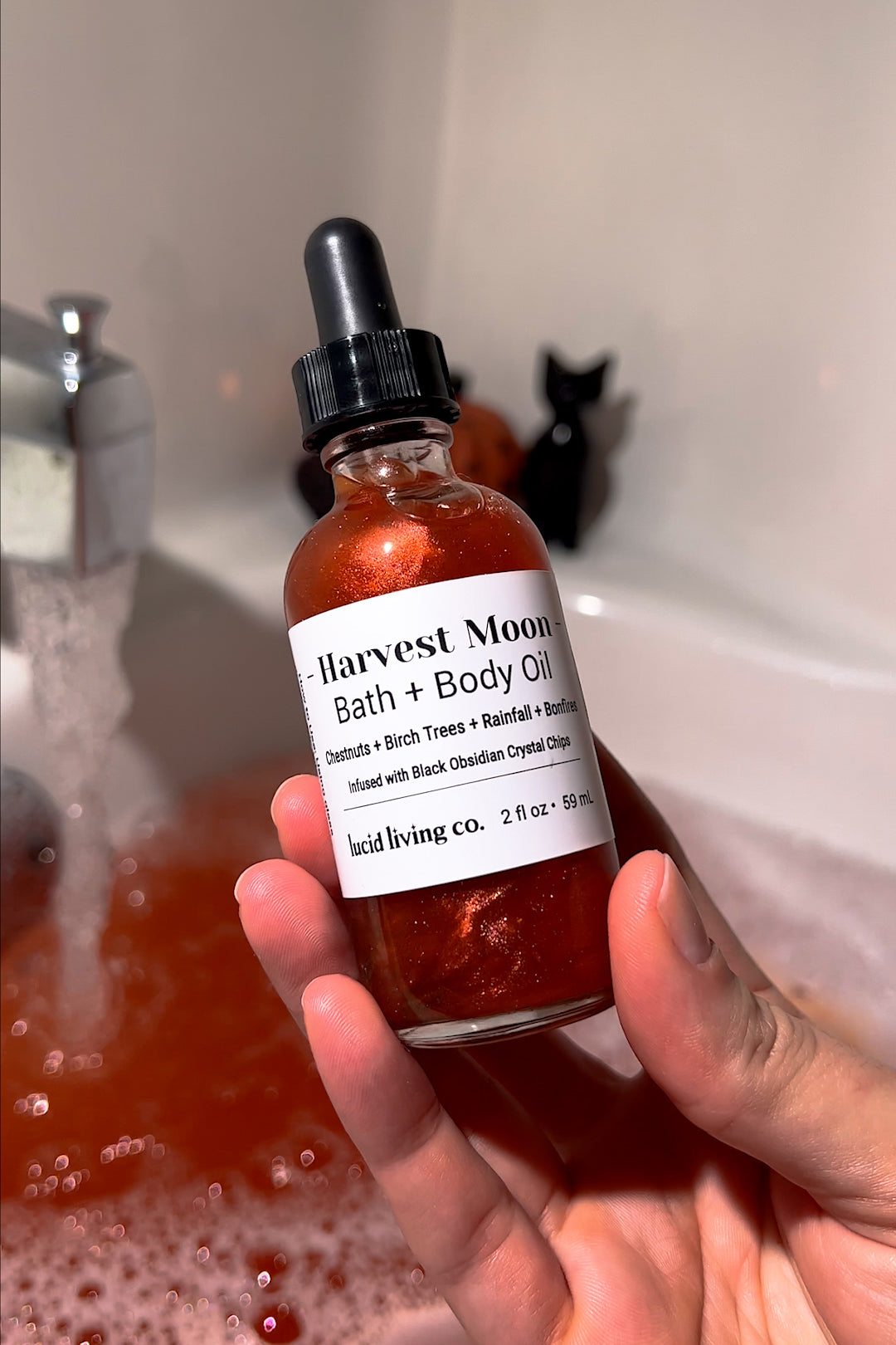 Hand holding Harvest Moon Body Oil bottle with shimmering liquid and black dropper over a bathtub background.