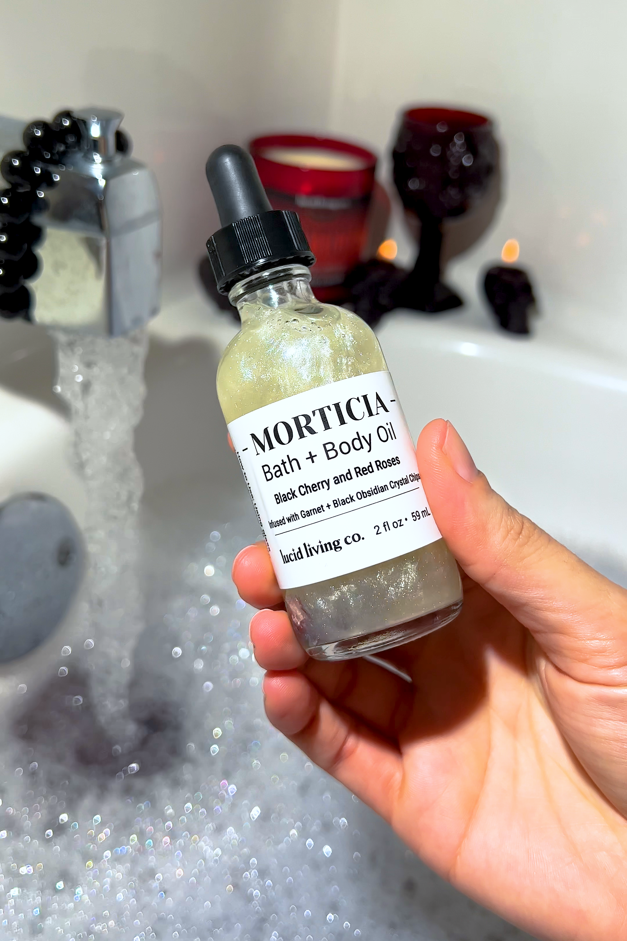 MORTICIA Bath &amp; Body Oil bottle with black cherry scent held over bubbly bathtub with dark candles nearby.