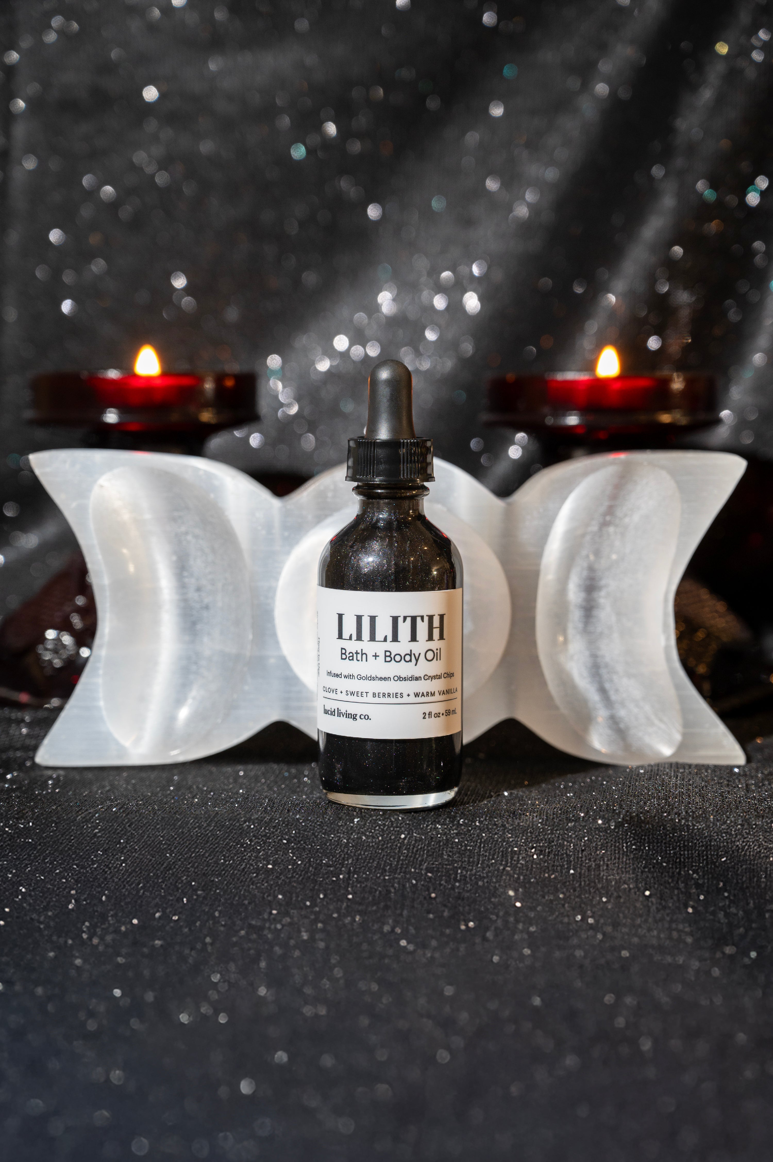Small bottle of LILITH Bath &amp; Body Oil with black dropper cap, positioned on a glittery dark surface between a white triple moon crystal and glowing red candles in the background.