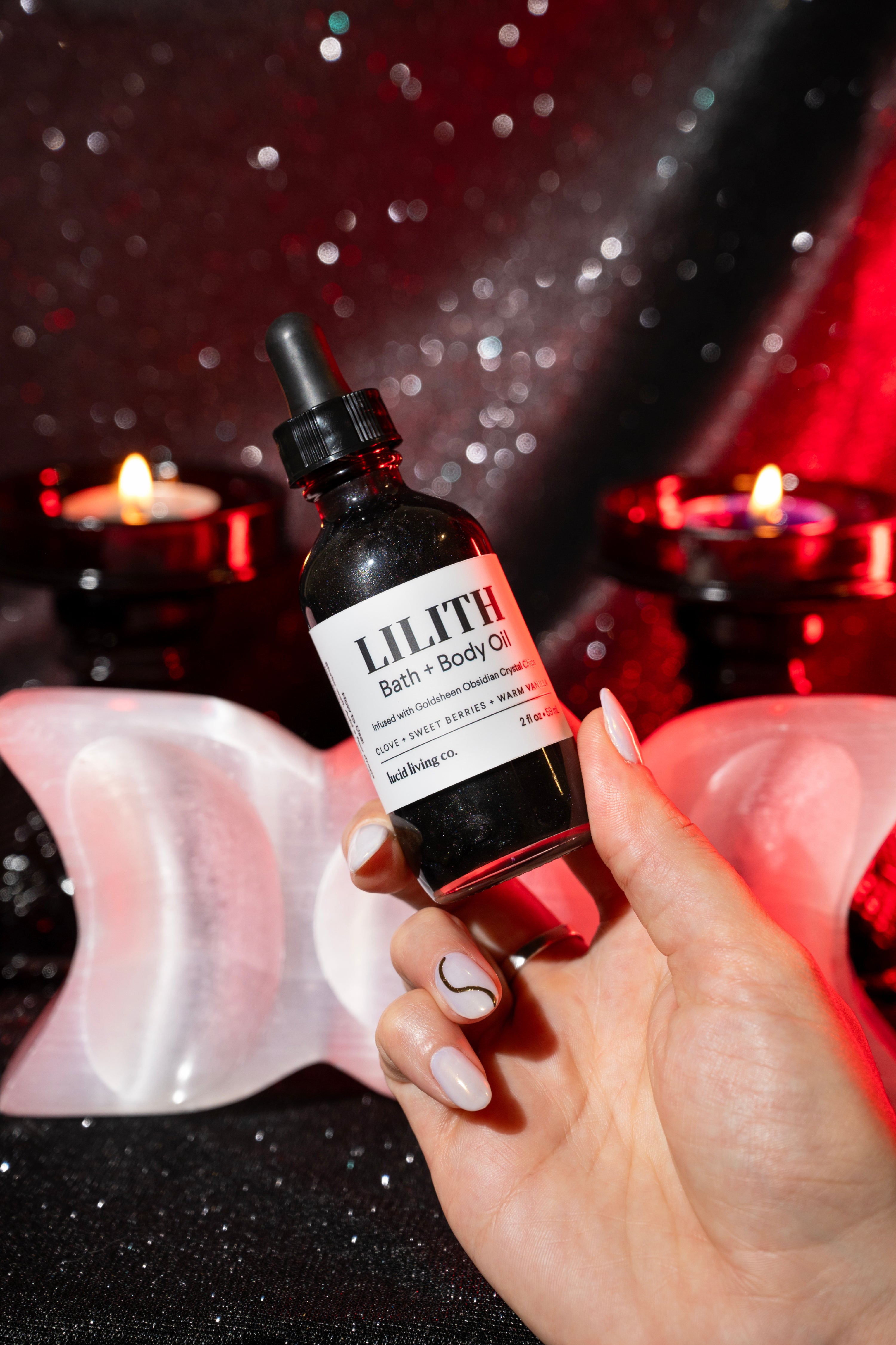 Hand holding a dark bottle of LILITH Bath &amp; Body Oil with a black dropper, glowing under red and white candlelight over a textured, sparkling black surface.