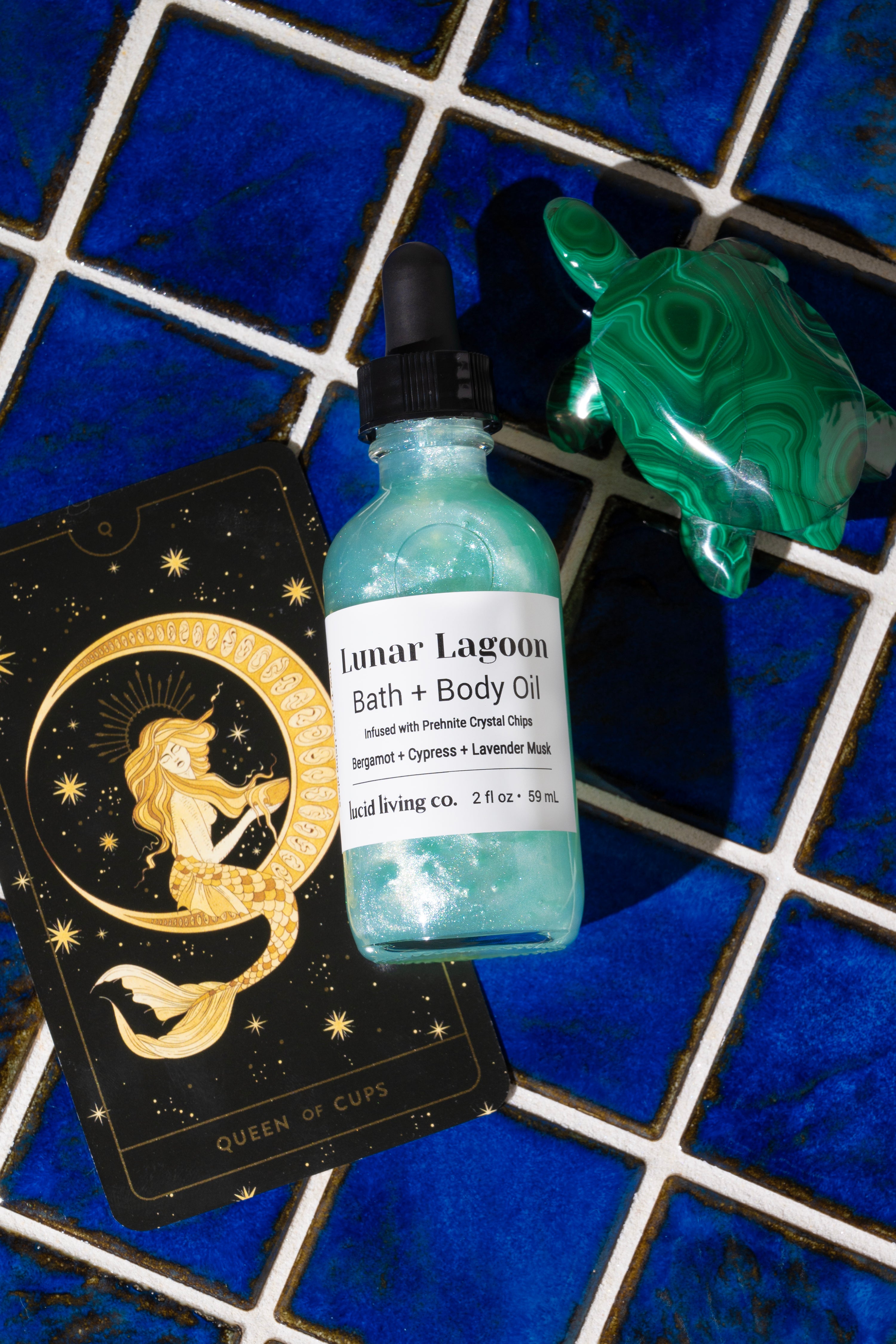 Lunar Lagoon Bath &amp; Body Oil bottle with a black dropper beside a green crystal turtle and an illustrated tarot card on blue tiled surface, showcasing a shimmering aqua color.