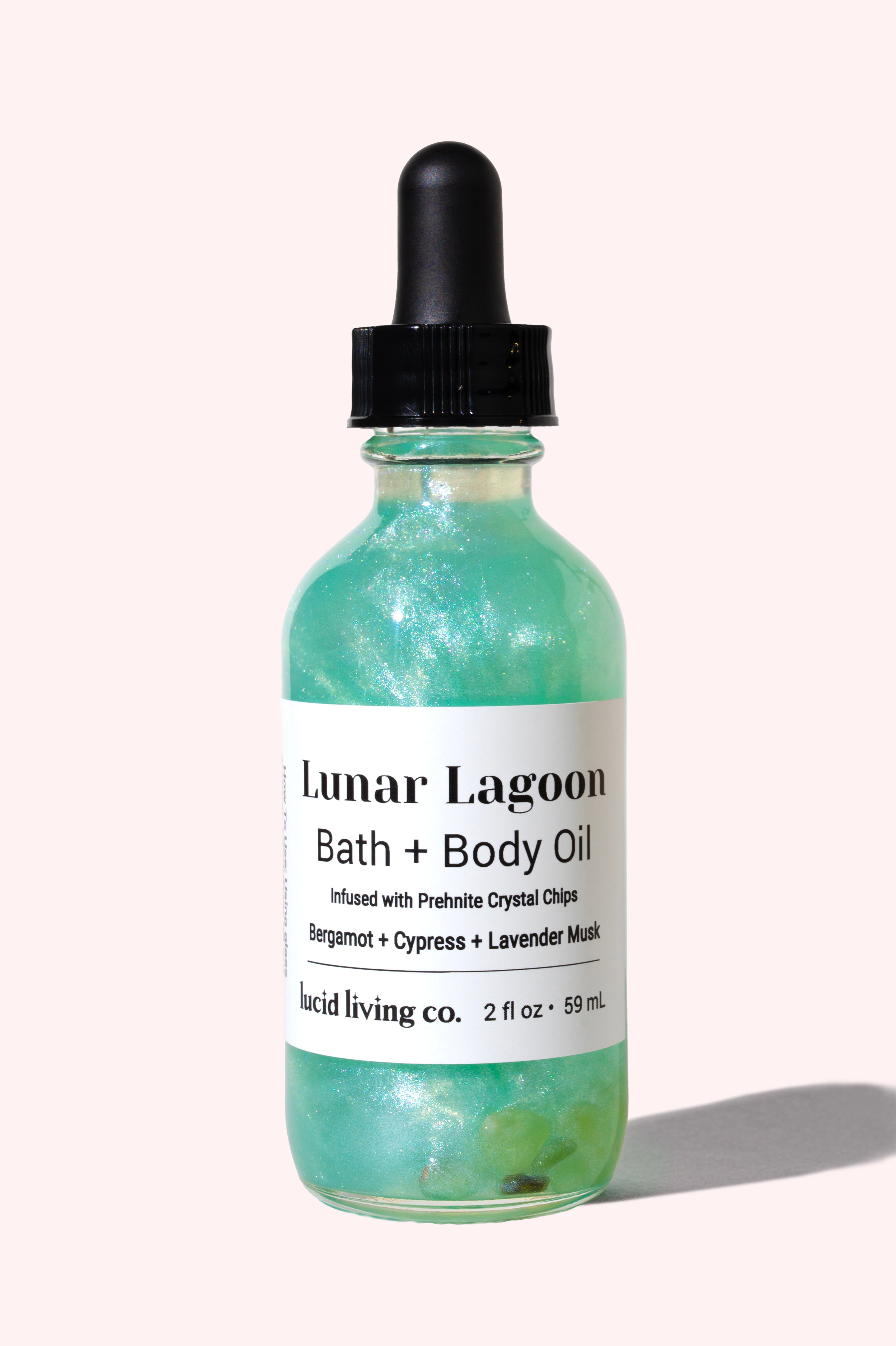 Clear glass bottle of Lunar Lagoon Bath &amp; Body Oil by Lucid Living Co., shimmering turquoise liquid with visible prehnite crystal chips inside and black dropper cap on pale background.